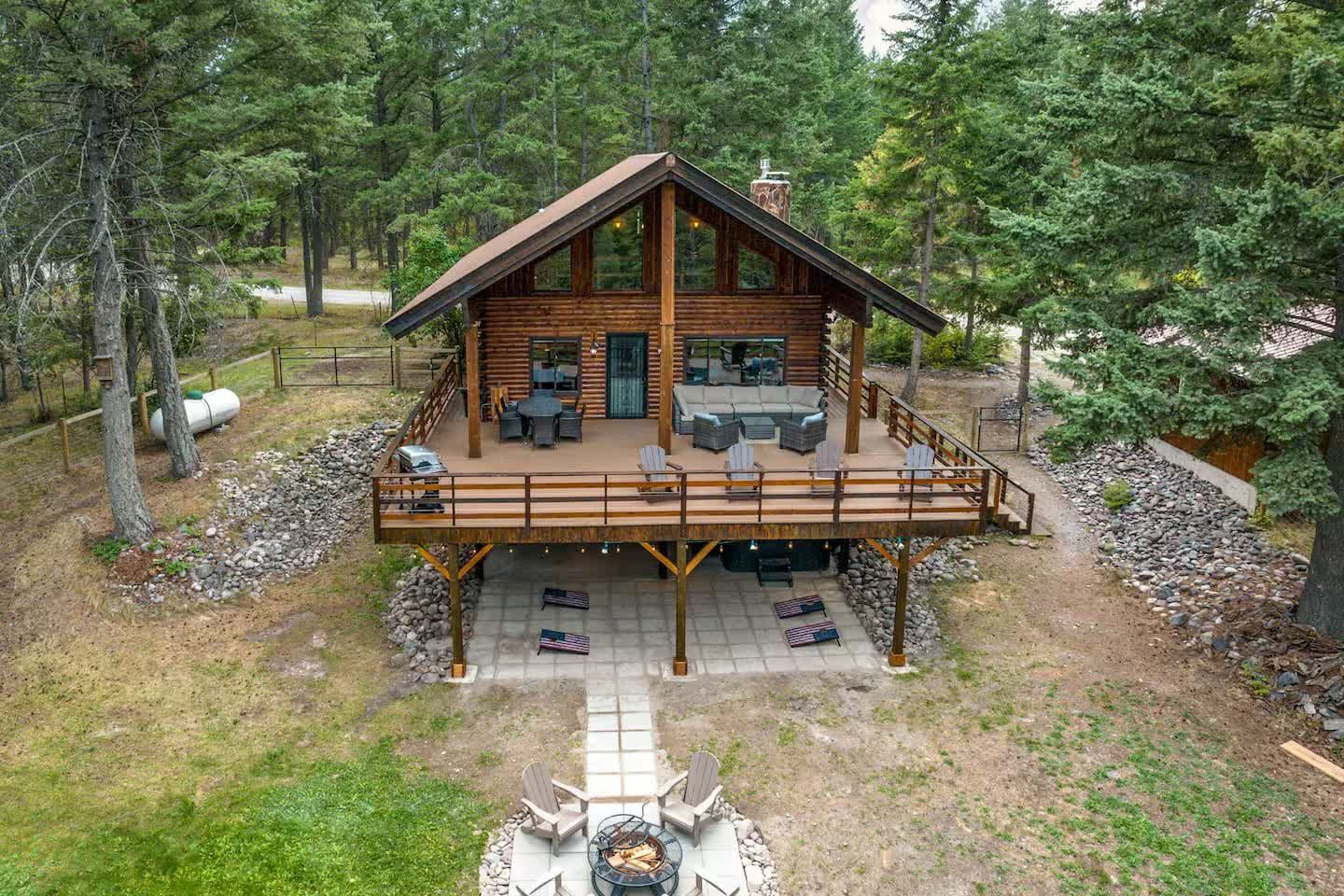 A log cabin in a forest with an elevated wooden deck, patio furniture, and a stone fire pit area on the ground below.