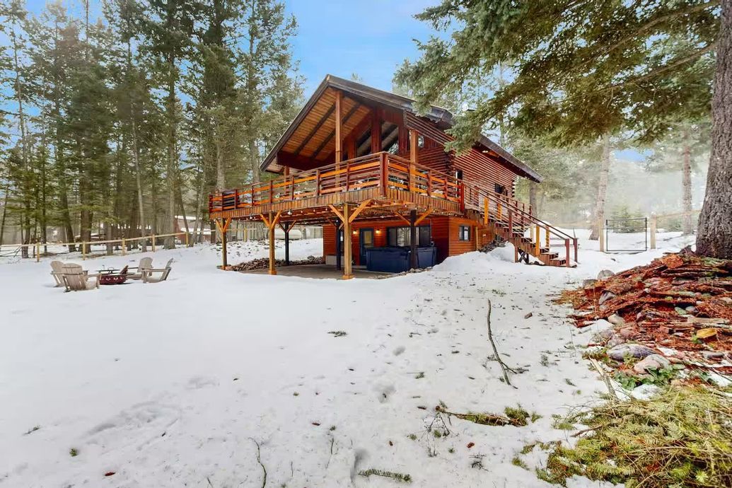 A wooden cabin with a large deck and ground-level hot tub nestled in a snowy, pine-covered forest.