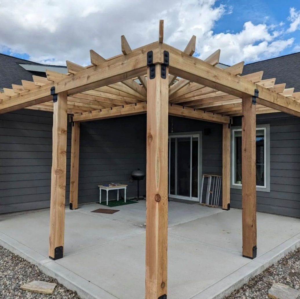 A wooden pergola with black metal brackets stands over a concrete patio next to a gray-sided house with a sliding door.