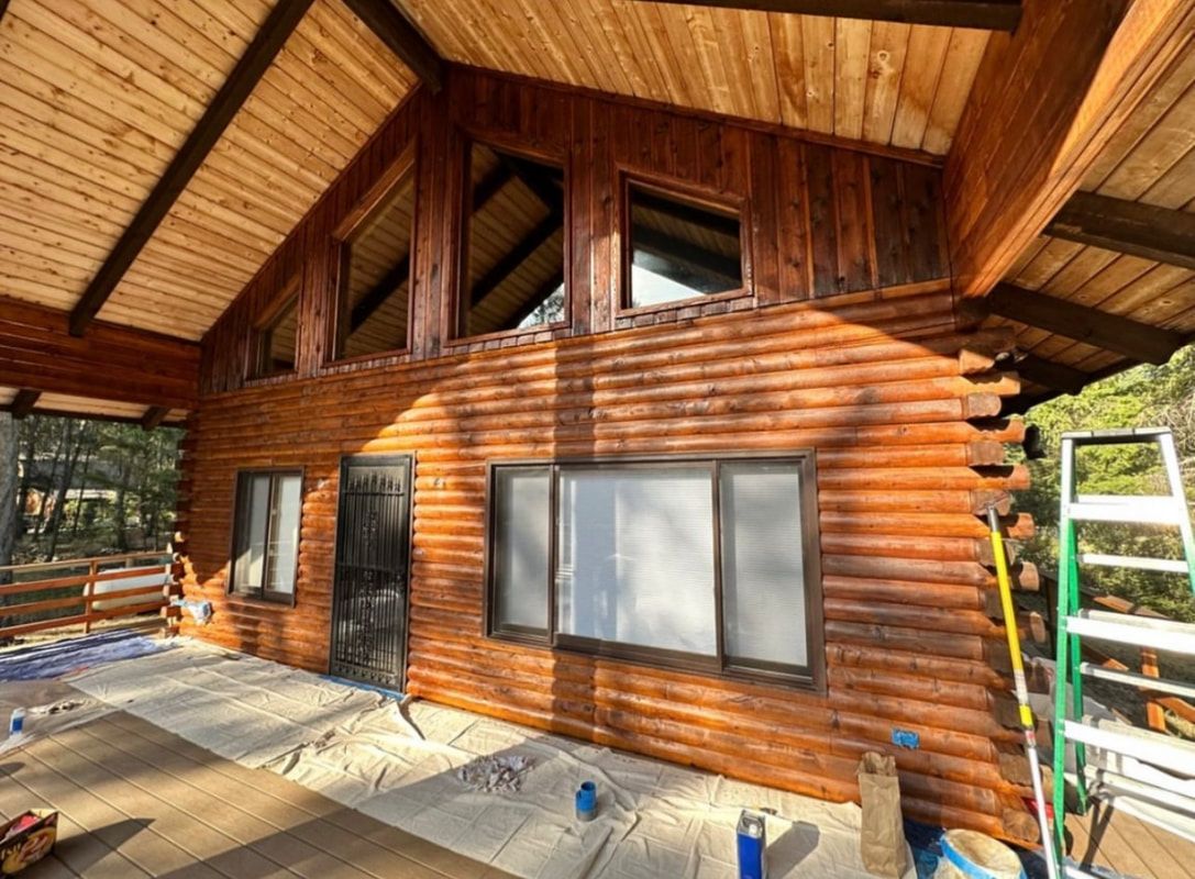 A rustic log cabin exterior with large windows, a wooden deck, and a ladder leaning against the side.