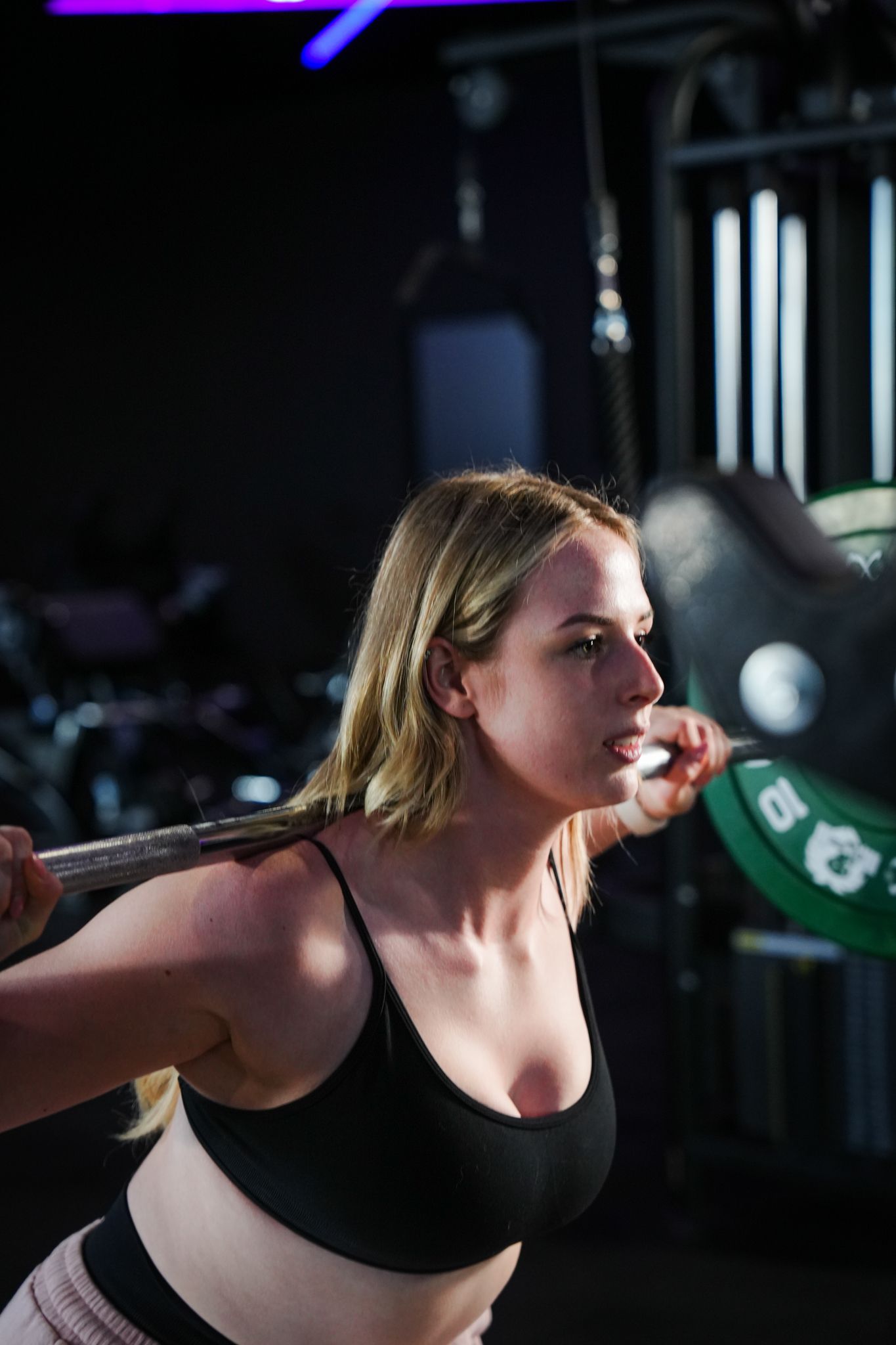Vrouw voert geconcentreerd en vastberaden squats uit met een halterstang in een sportschool. Zwarte sportbeha, gewichten aan de stang.