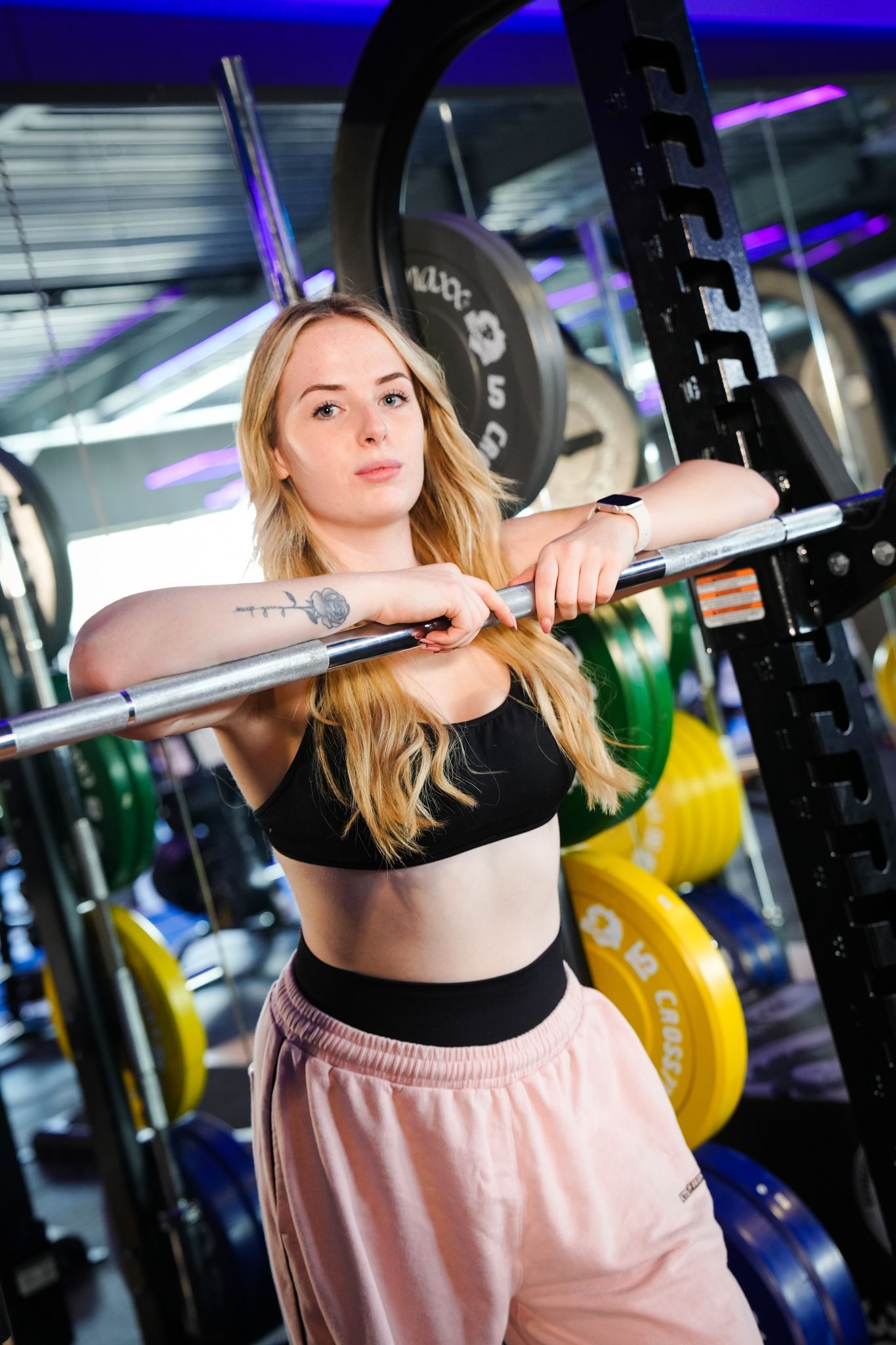 Vrouw in een sportschool, leunend op een halter, gekleed in een zwarte crop top en roze joggingbroek.