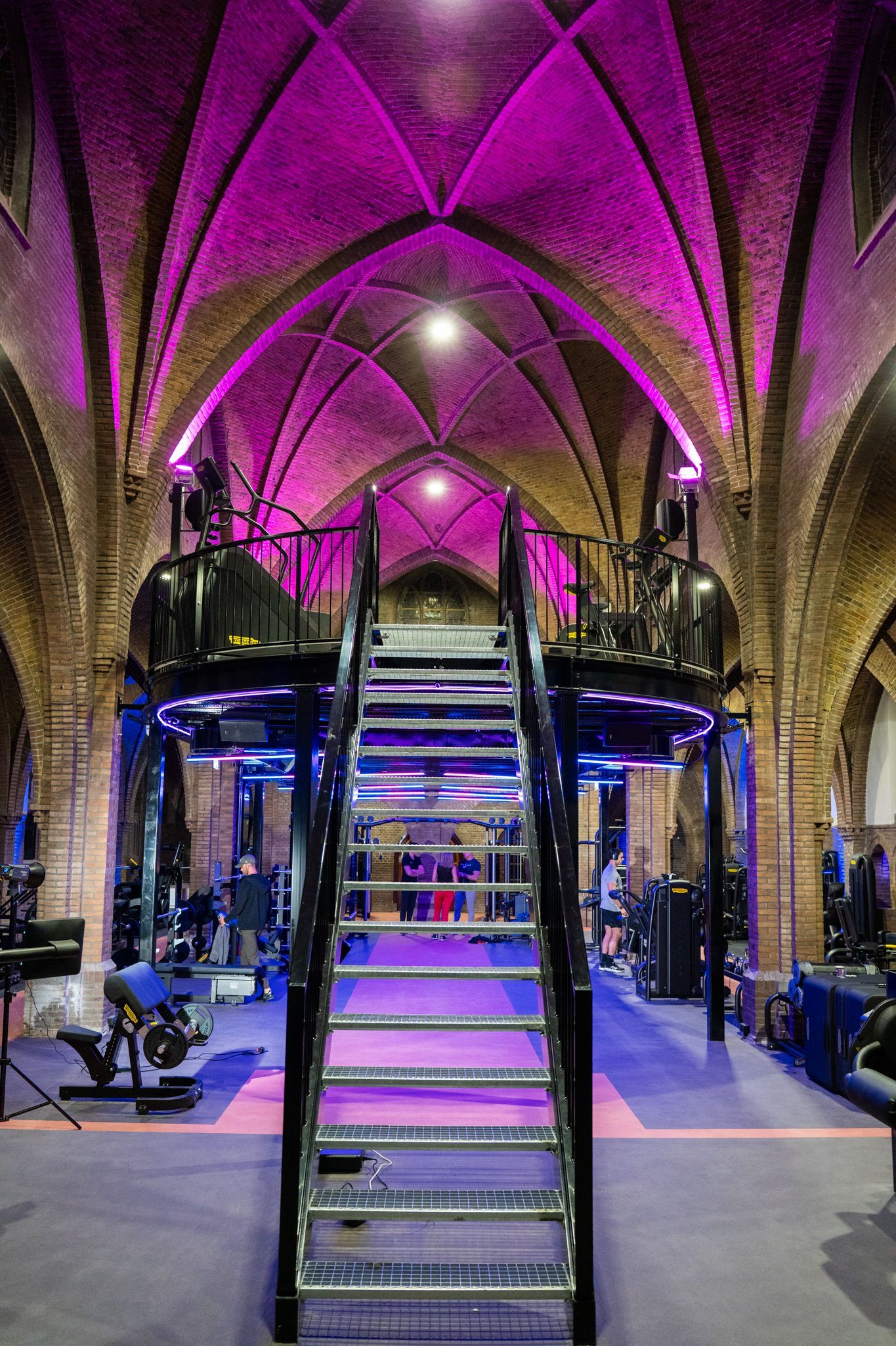 Sportschoolinterieur met gewelfd plafond en roze verlichting. Een metalen platform en een trap bevinden zich in het midden.