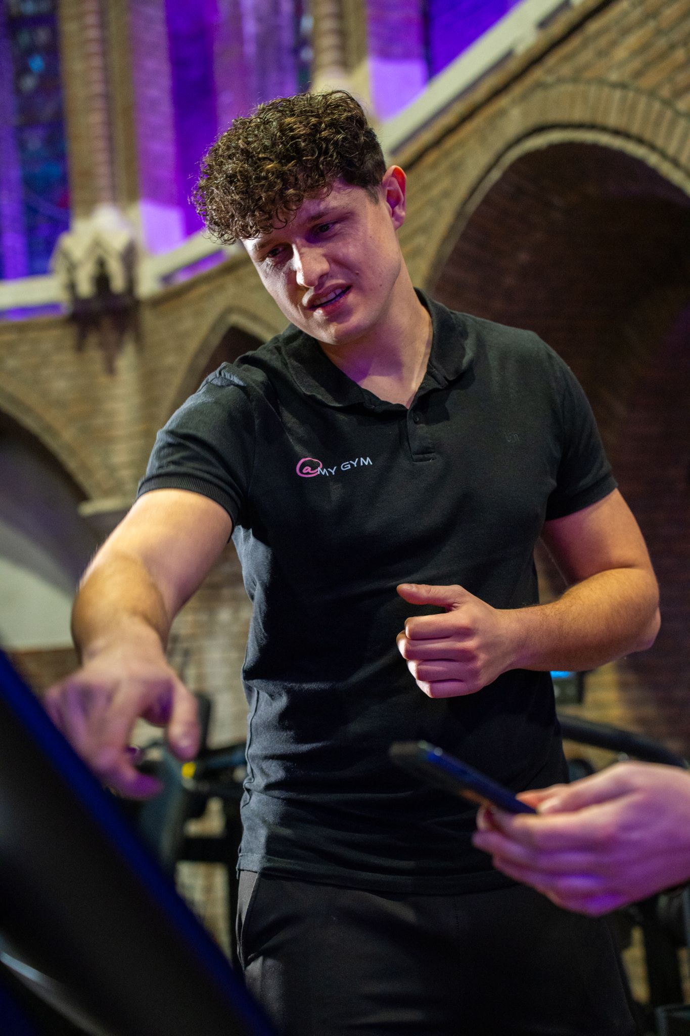 Een man in een zwart poloshirt wijst naar een loopband en praat met iemand die een telefoon vasthoudt. Paarse verlichting op de achtergrond.