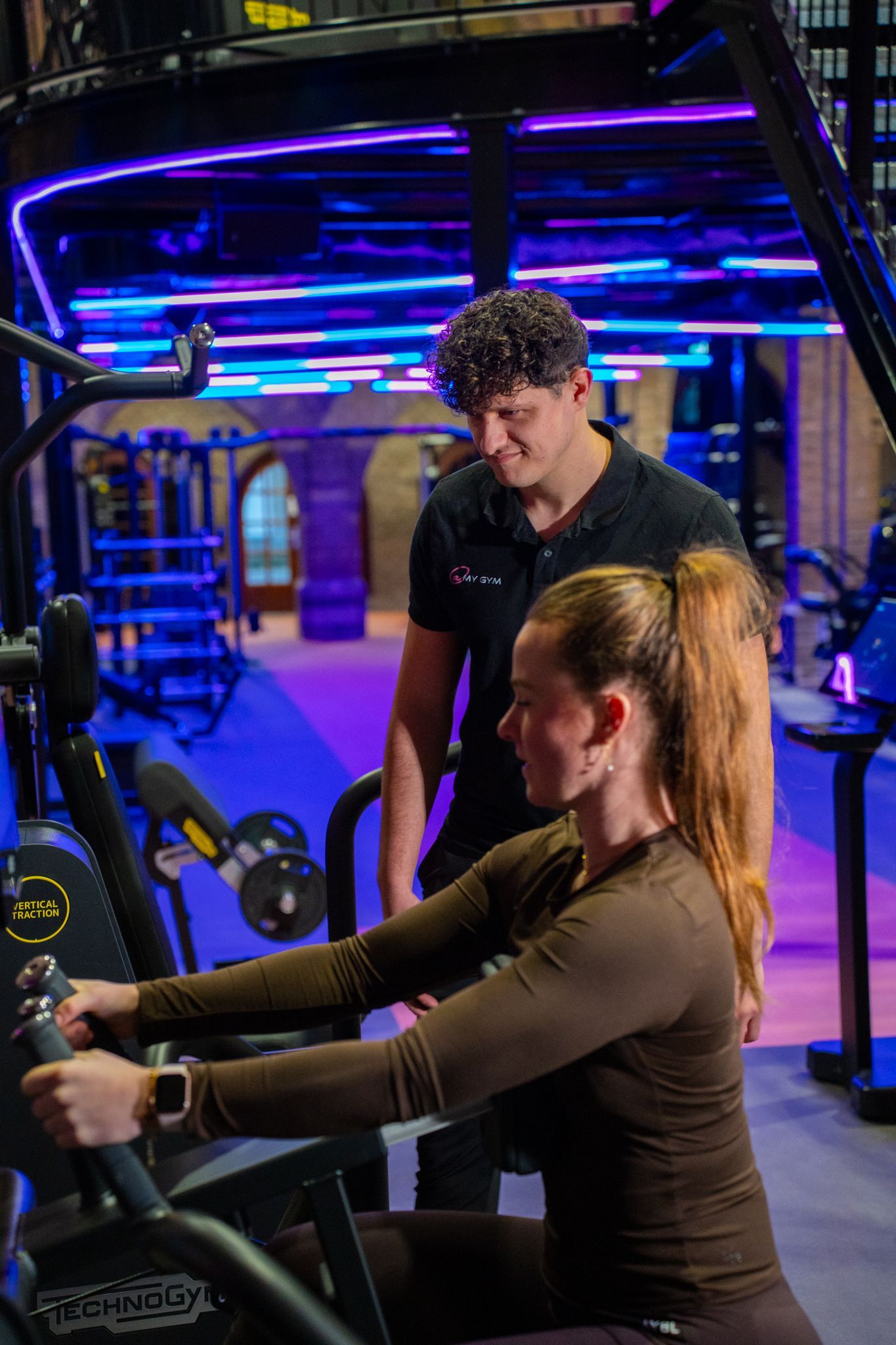 Een vrouw gebruikt een roeimachine, terwijl een trainer toekijkt in een sportschool. Paarse en blauwe verlichting.