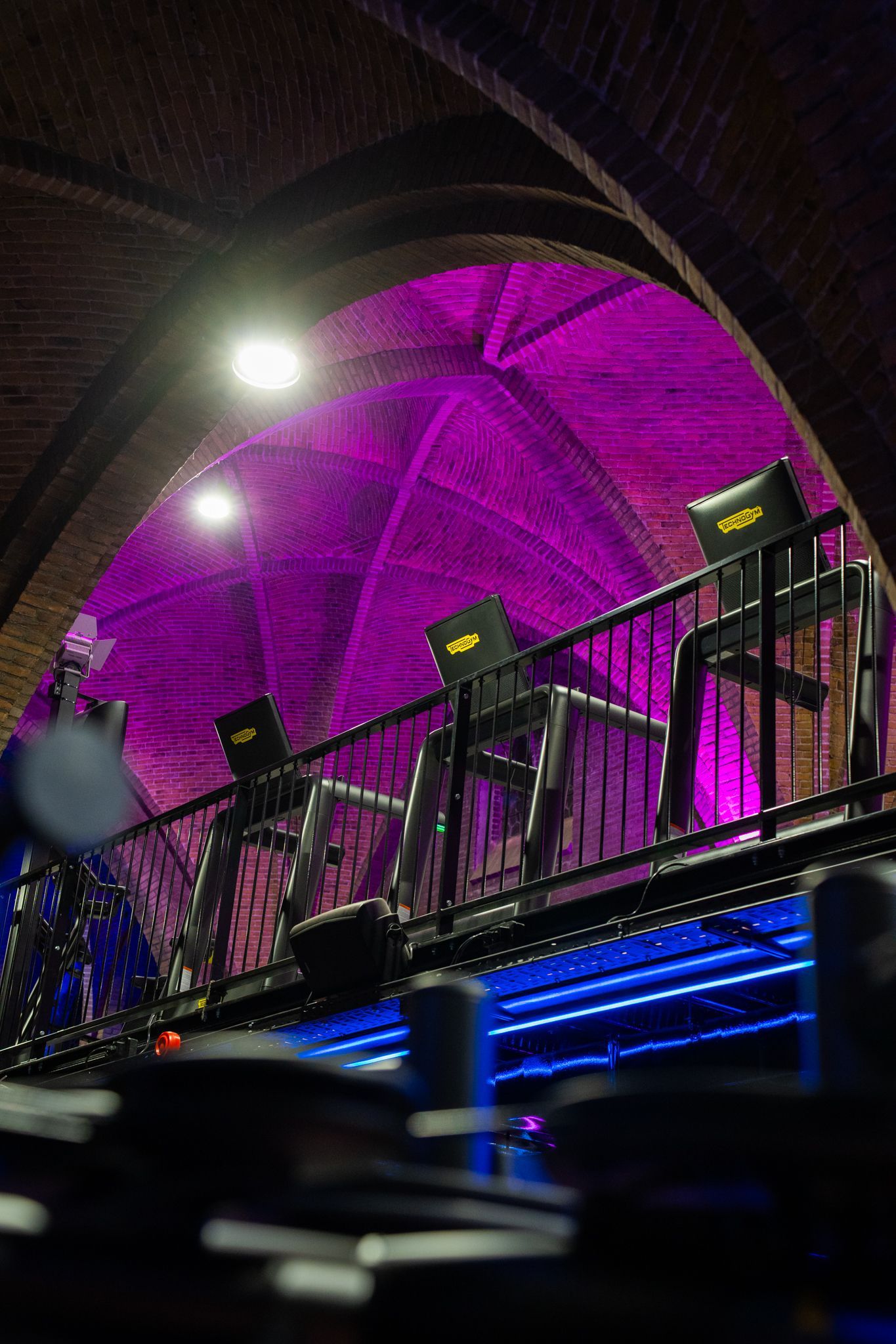 Drie modern ogende stoelen op een balkon met paarse verlichting in een ruimte met een bakstenen boog.