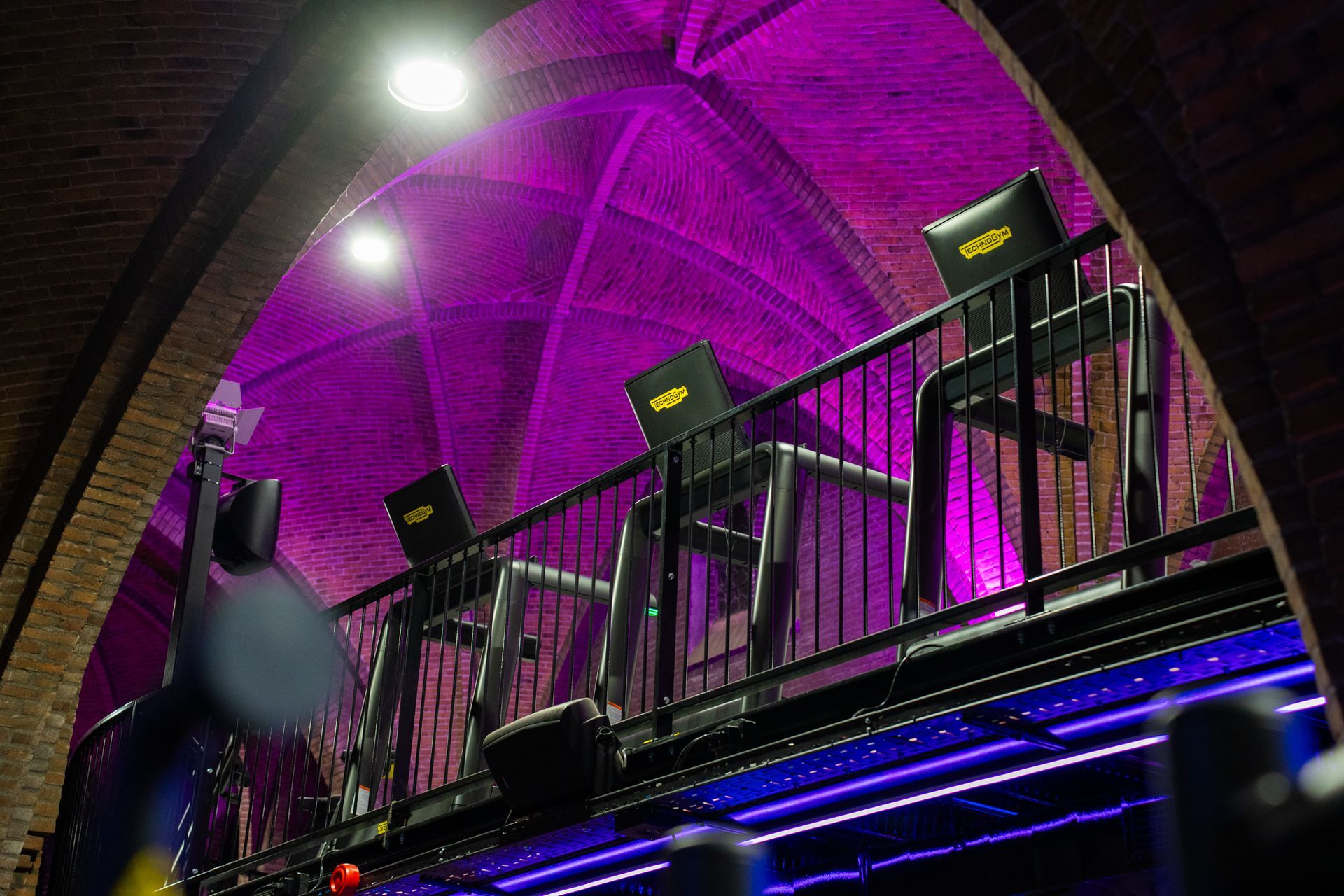 Fitnessapparatuur op een balkon met een gewelfd plafond en paarse verlichting.