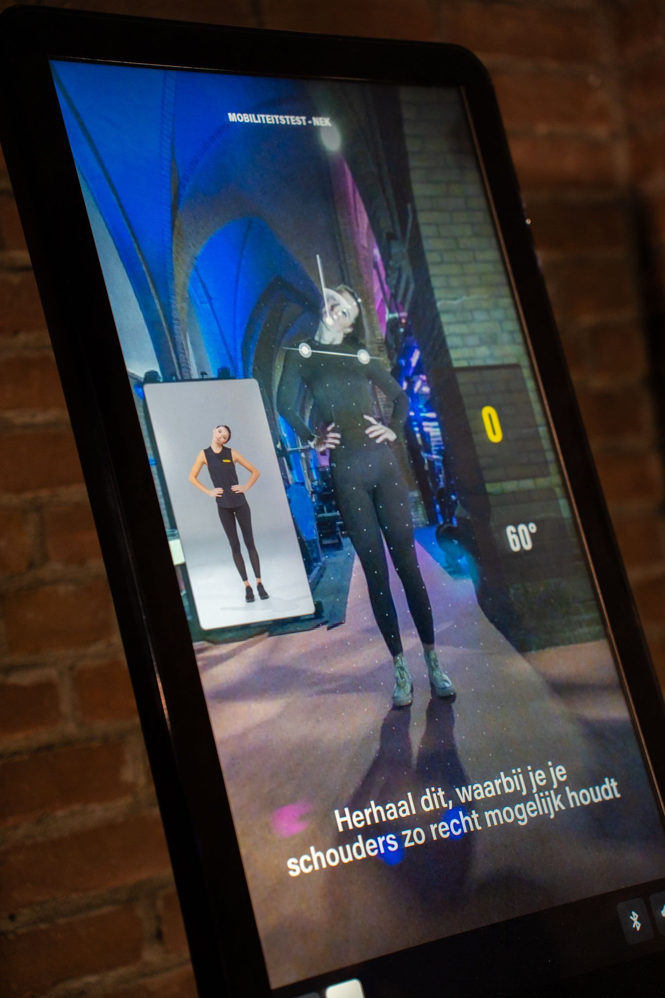 Op een tablet wordt een virtuele fitnessinstructeur in een studio-omgeving weergegeven. Een detailweergave toont een voorbeeldoefening.