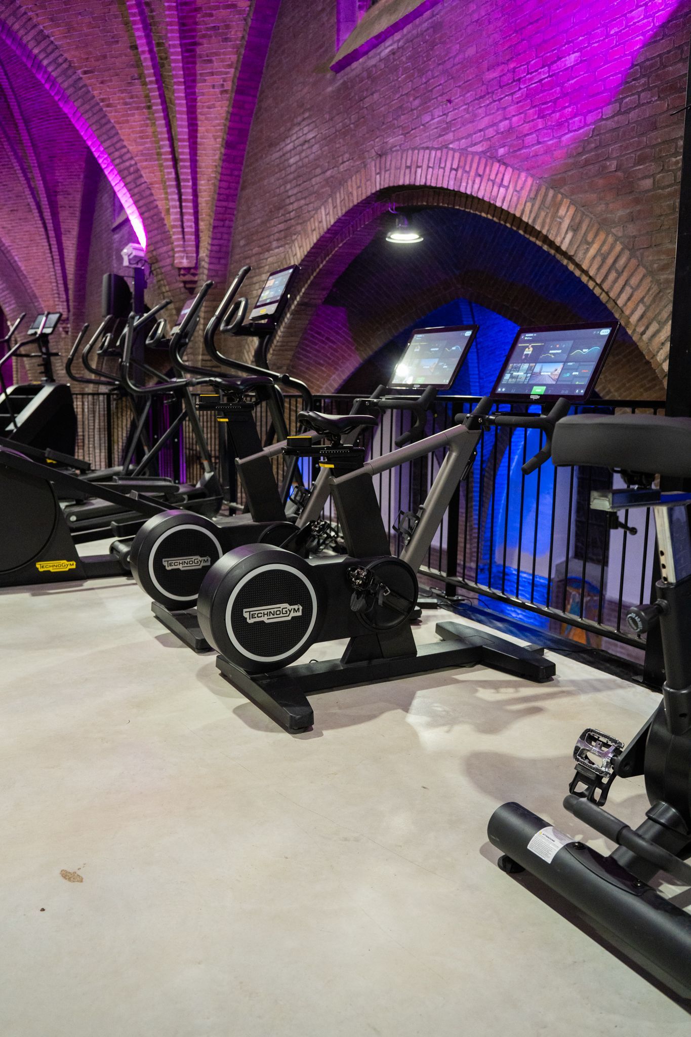 Interieur van een sportschool met fitnessapparaten onder een gewelfd bakstenen plafond, verlicht met paars licht.