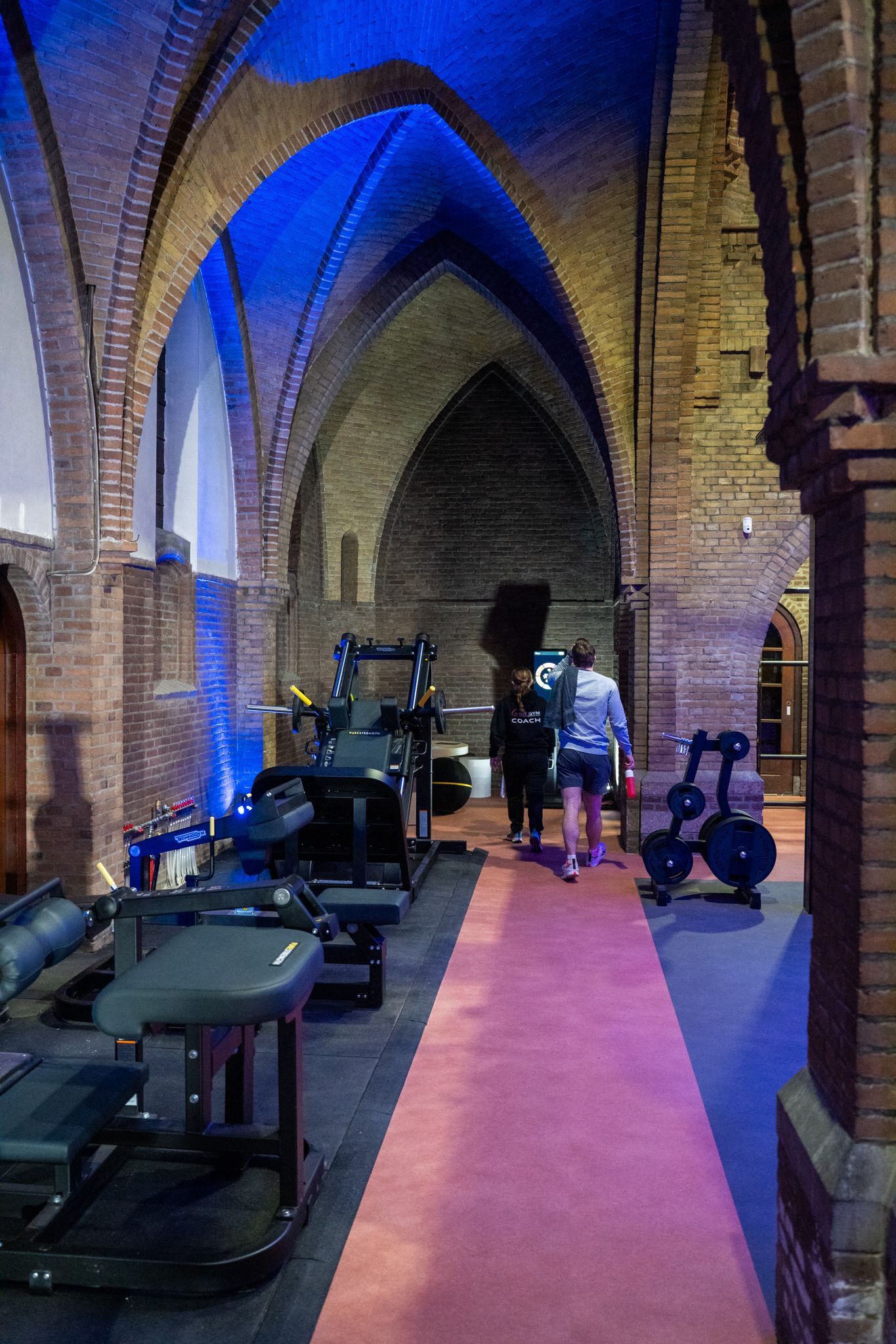 Fitnessapparatuur in een gewelfd bakstenen gebouw met een blauw verlicht plafond; twee mensen lopen over een rode loper.