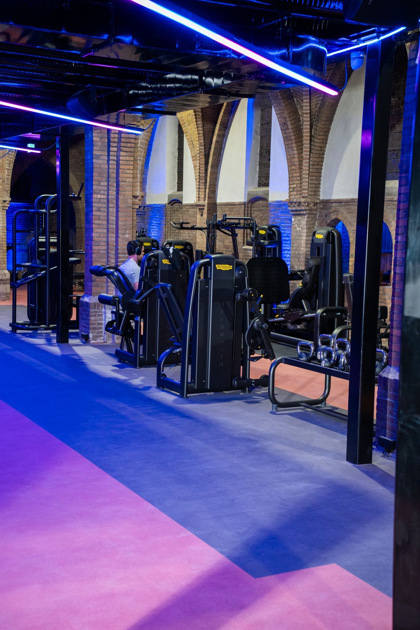 Interieur van een sportschool met fitnessapparaten tegen bakstenen bogen, verlicht door paarse en blauwe neonlichten.