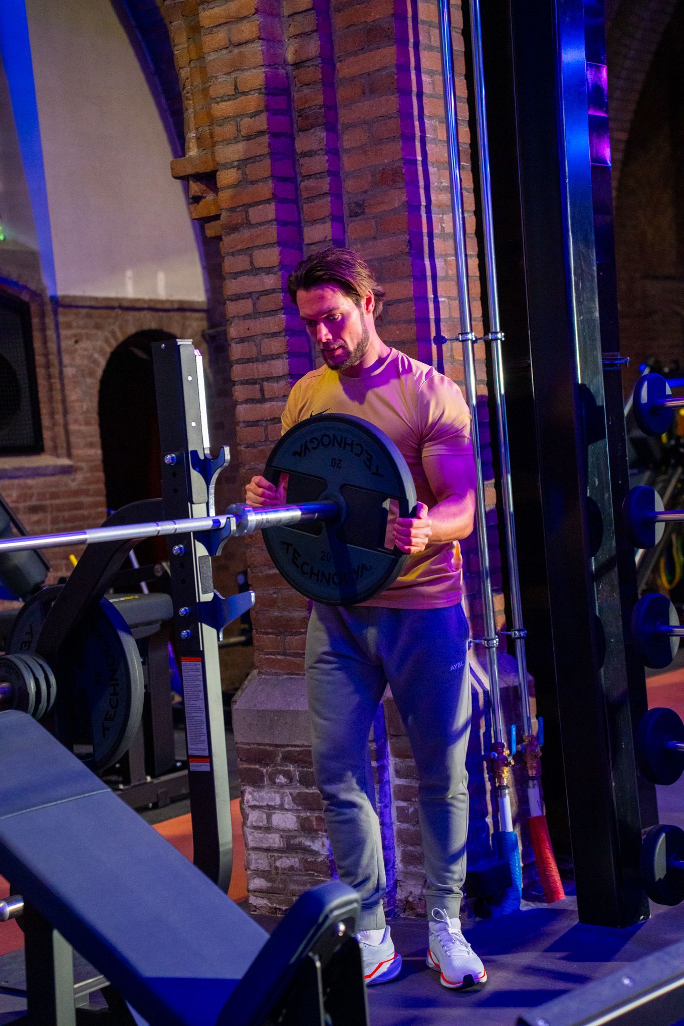 Een man voegt een gewichtsschijf toe aan een halterstang op een gewichtsbank in een sportschool met paarse en blauwe verlichting.