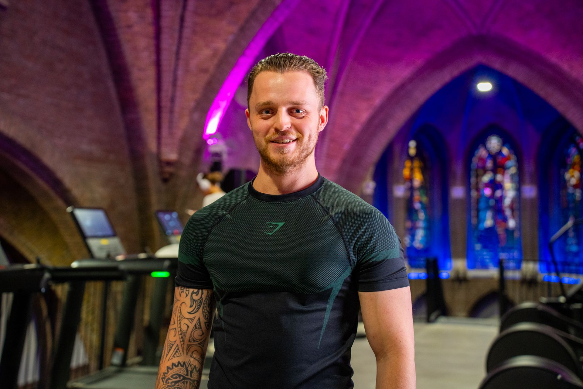 Een man in sportkleding lacht in een sportschool met een gewelfd plafond en glas-in-loodramen; op de voorgrond is gewichtsapparatuur te zien.