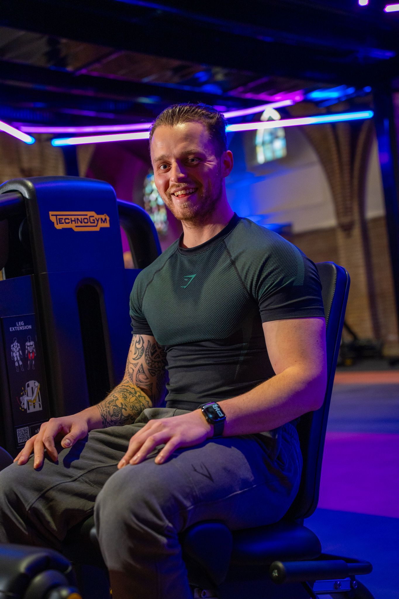 Een man zit lachend op een fitnessapparaat in een sportschool met kleurrijke verlichting.
