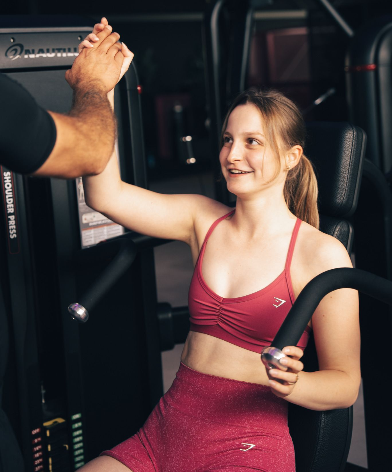 Een vrouw in rode sportkleding geeft een high-five aan een trainer in de sportschool na het gebruik van een schouderpers.