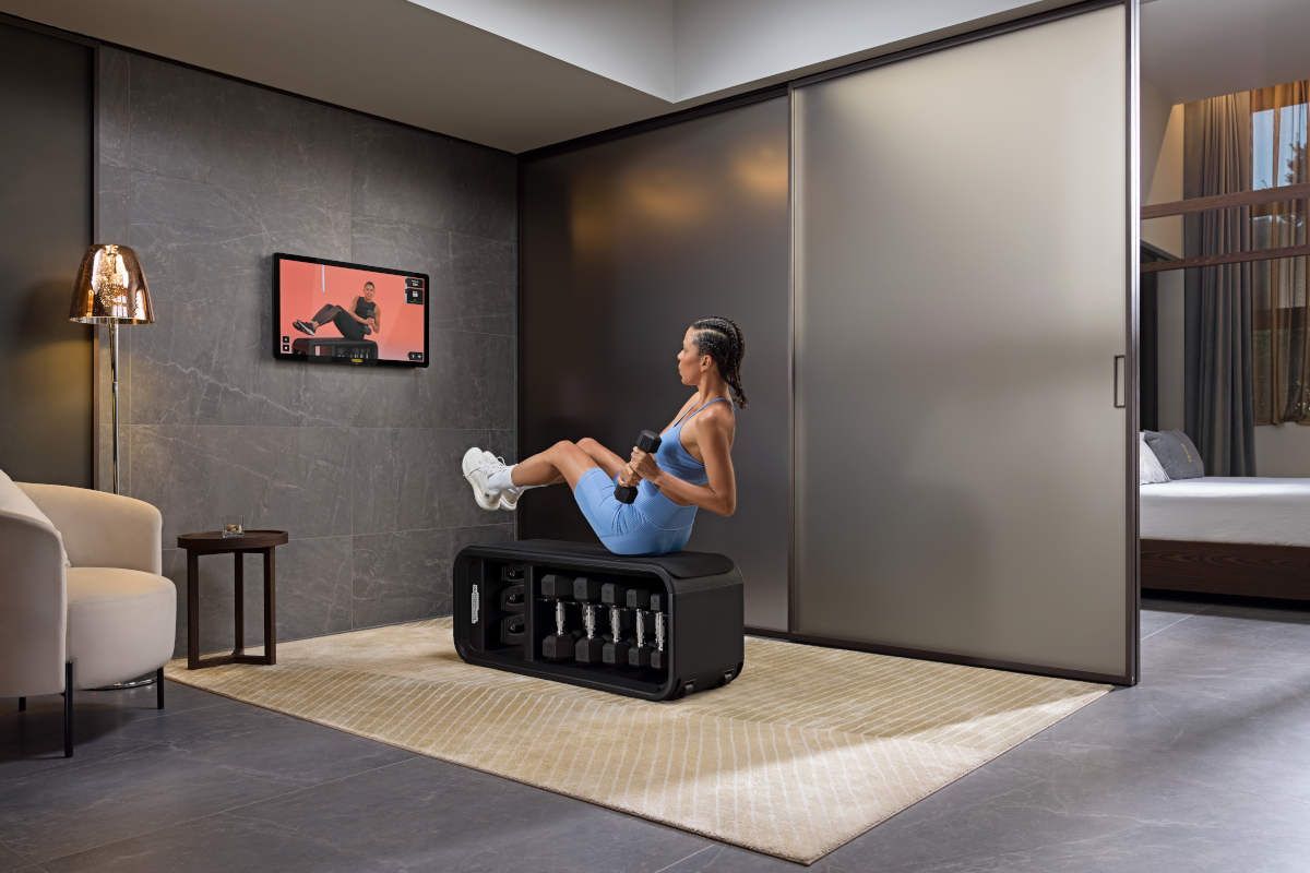 Een vrouw doet oefeningen met gewichten voor een scherm waarop een trainingsprogramma wordt weergegeven, in een moderne kamer.