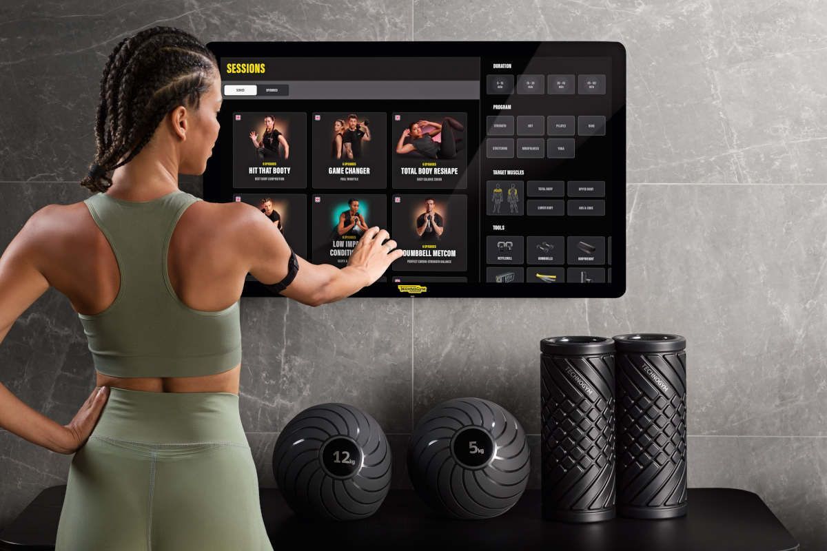 Vrouw gebruikt een fitnessconsole met touchscreen waarop trainingsvideo's worden bekeken; fitnessapparatuur is in de buurt.