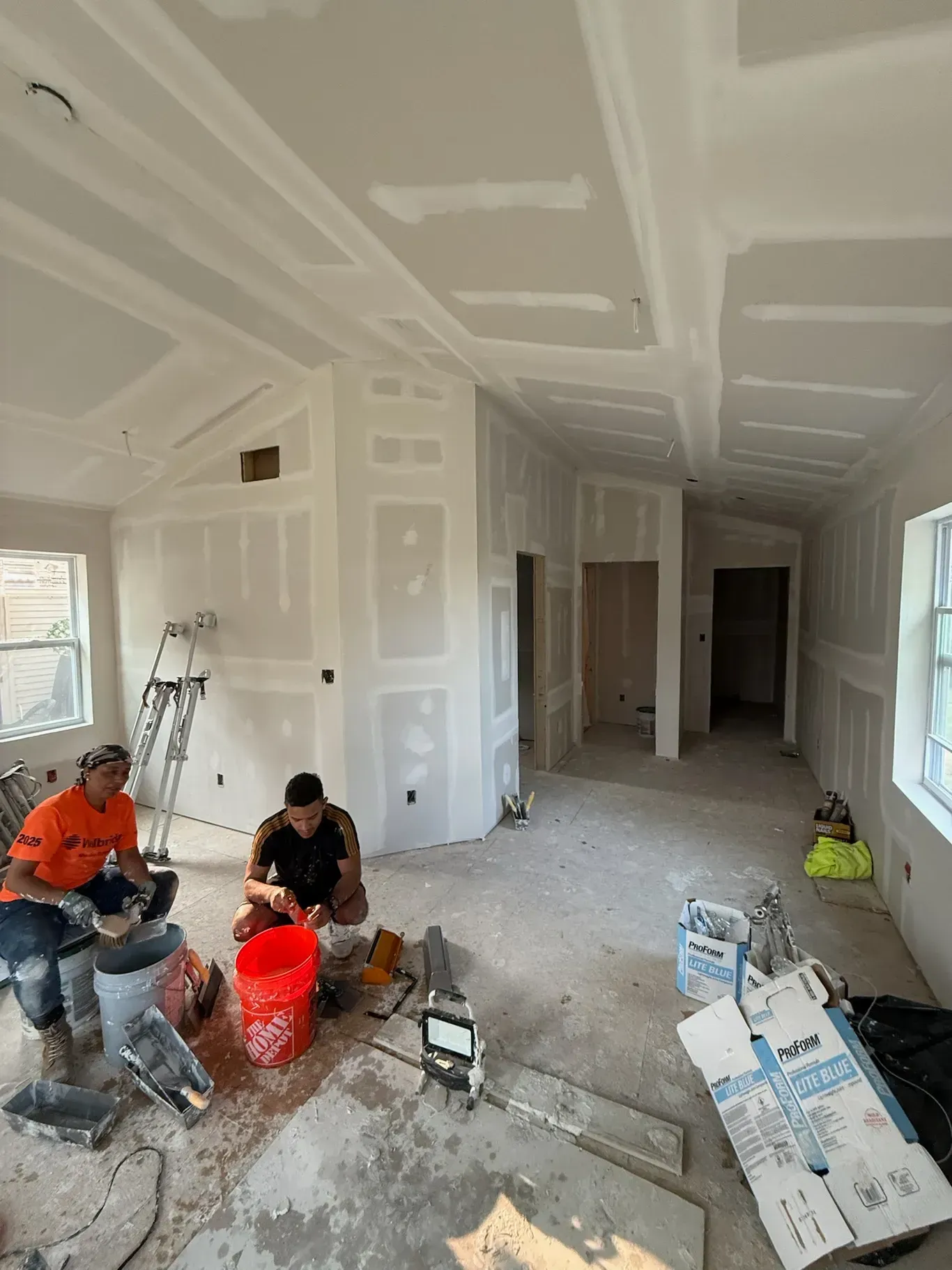 Construction workers installing drywall inside a building. One is mixing mud, another is watching.