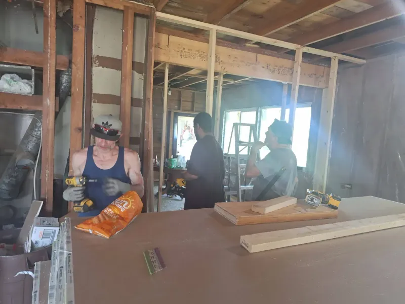 Construction workers inside a building. One uses a drill; others observe near a window. Unfinished walls and debris are visible.