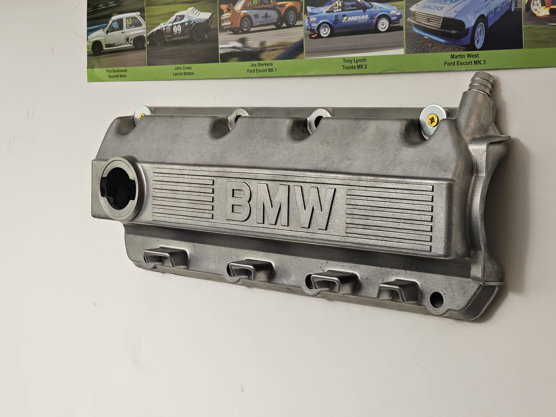 Silver BMW engine valve cover mounted on a white wall with racing cars in the background.