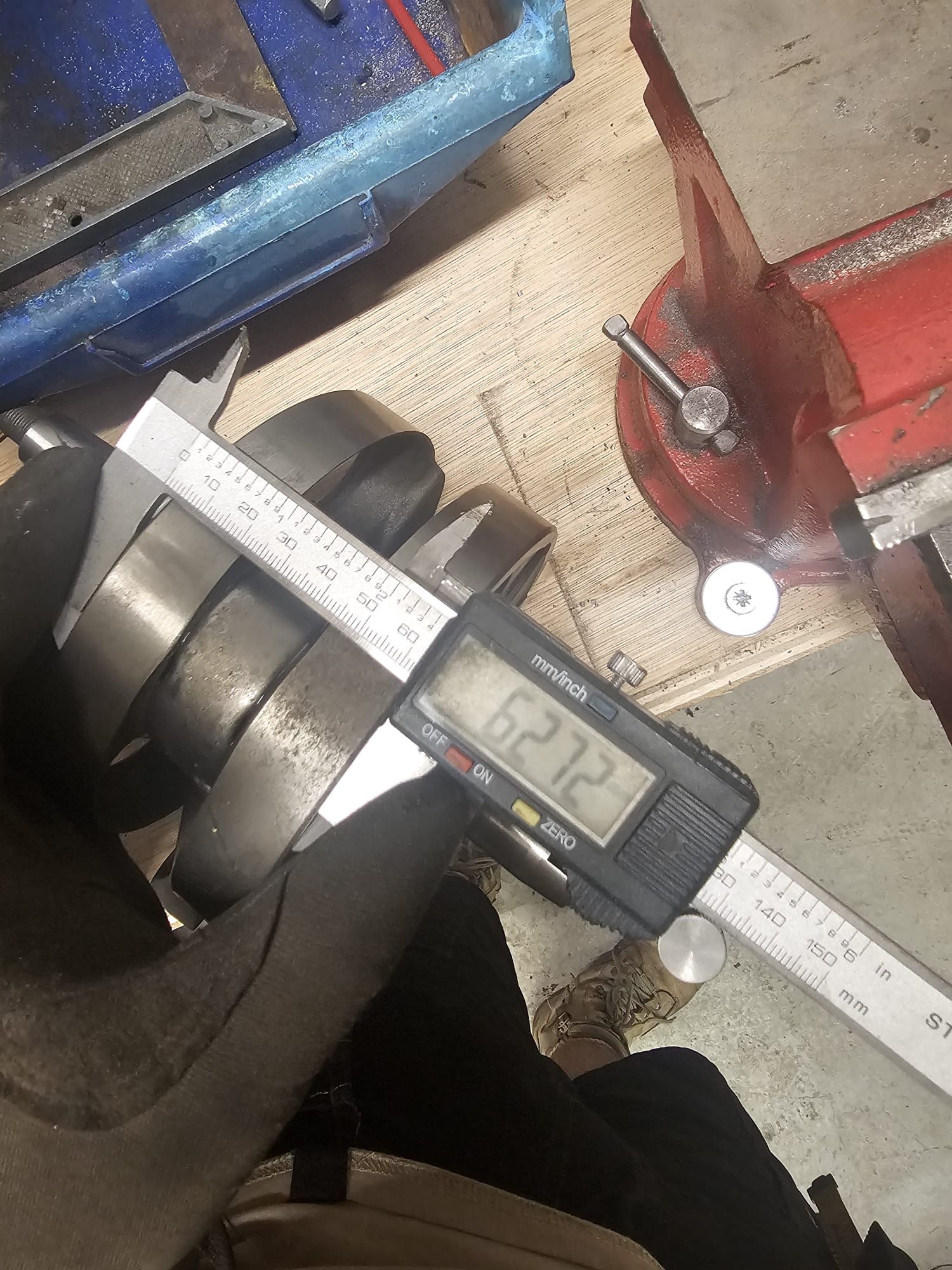 A person measures the diameter of a cylindrical metal object with digital calipers in a workshop. The reading is 62.72.