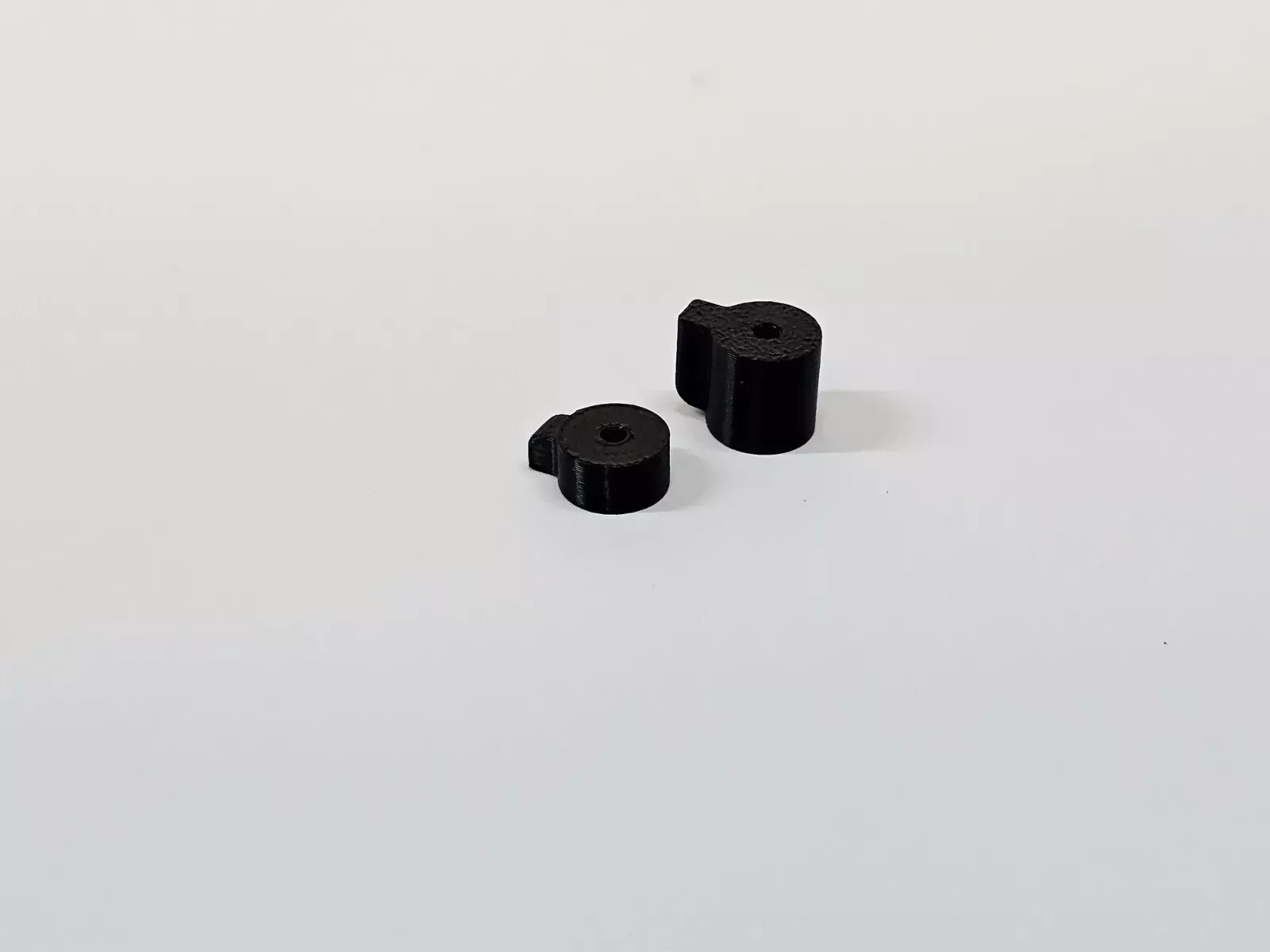 Two small, black, cylindrical 3D-printed objects on a white surface. Each has a small notch and a central hole.