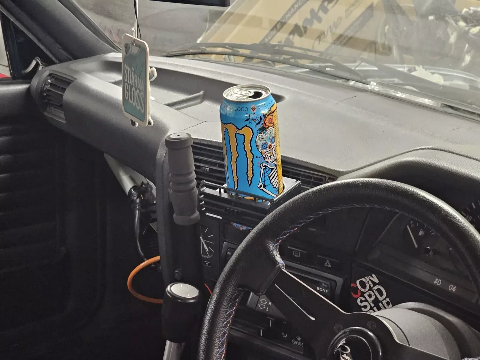 A can of Monster energy drink sits on the dashboard of a classic car, near the steering wheel and a shifter.