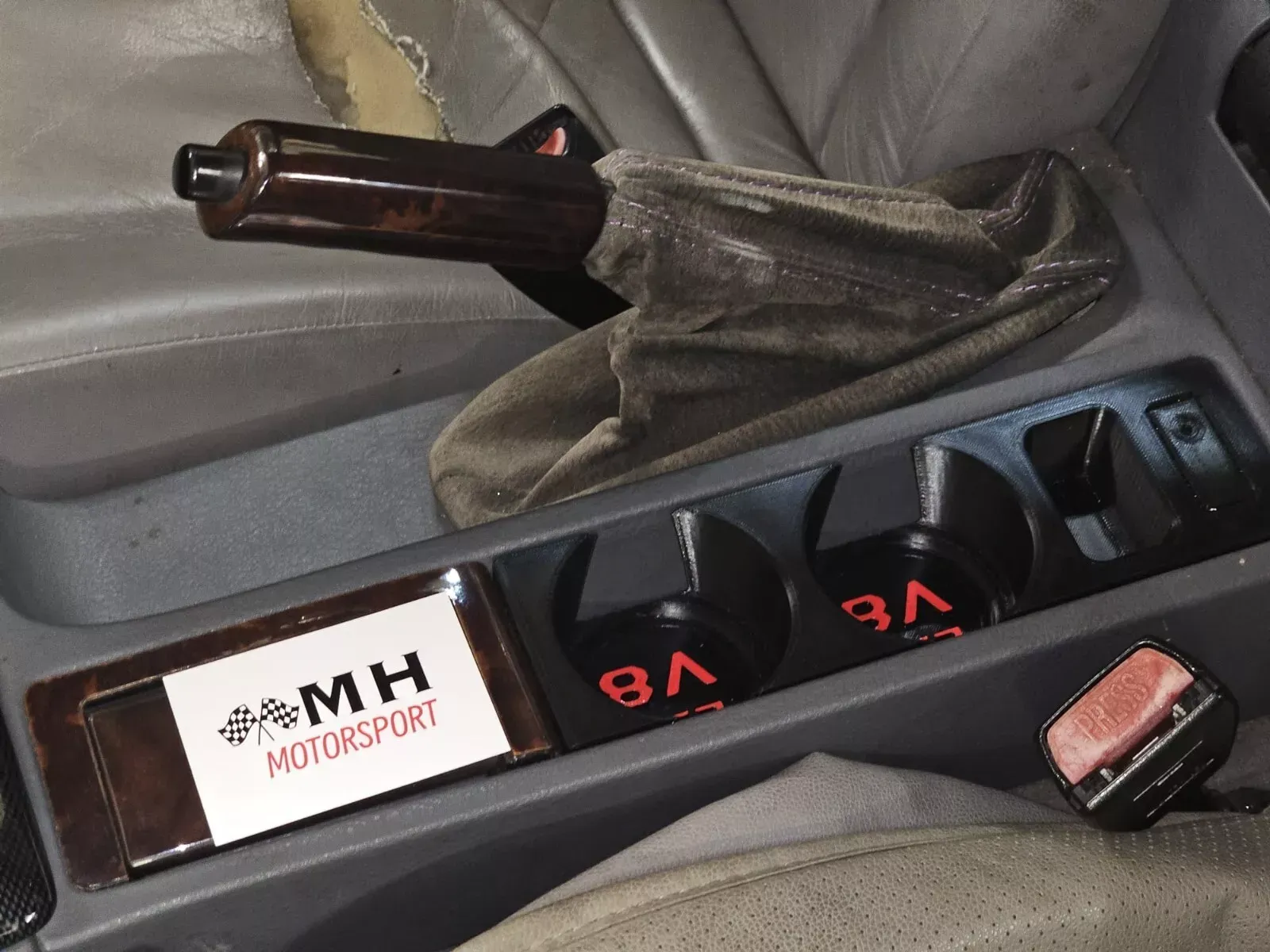 Car interior: center console with cup holders, emergency brake, V8 logo, and MH Motorsport card.