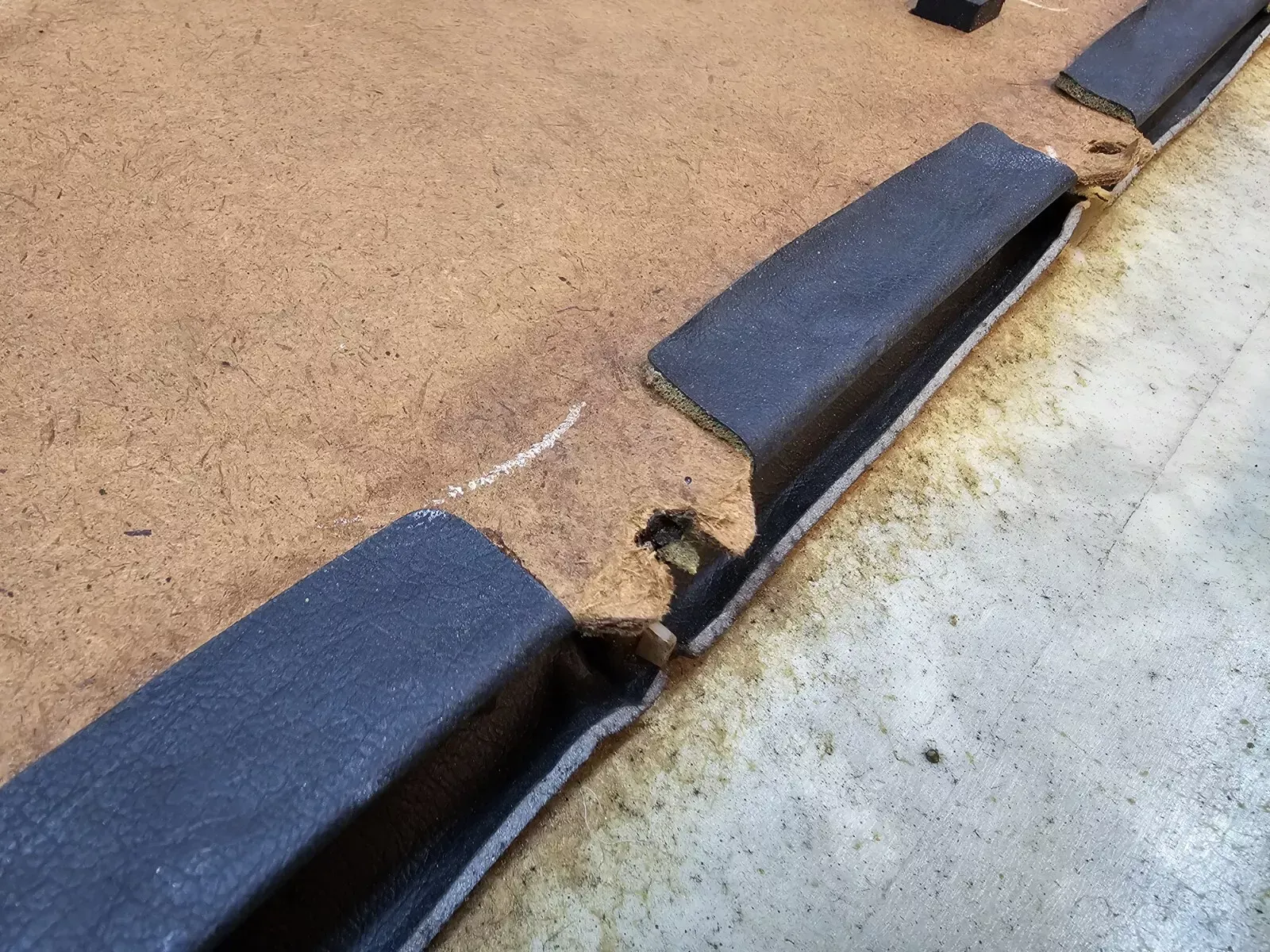 Damaged dark molding on a brown panel with exposed beige underlayer, against a light grey surface.