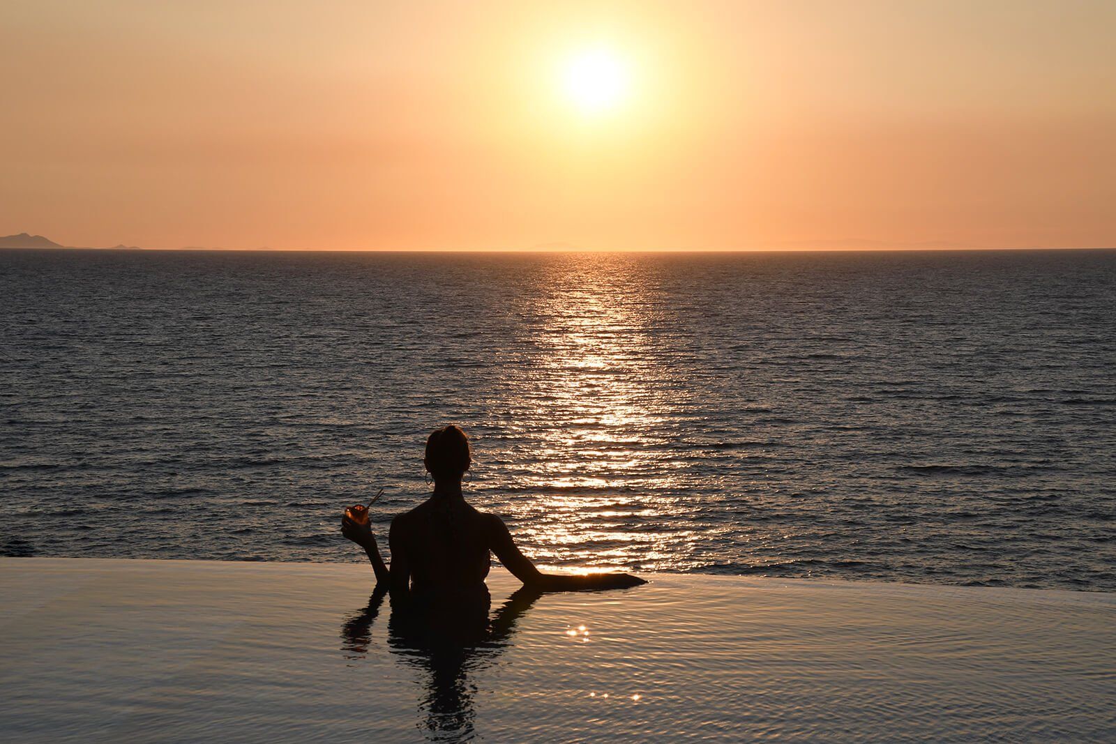 A woman is sitting in a pool watching the sun set over the ocean