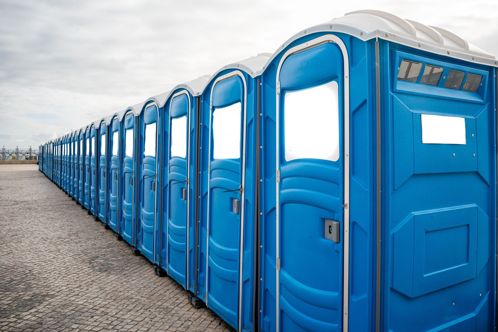 Long row of blue portable toilets with white roofs on a paved outdoor area. Long row of blue portable toilets with white roofs on a paved outdoor area.