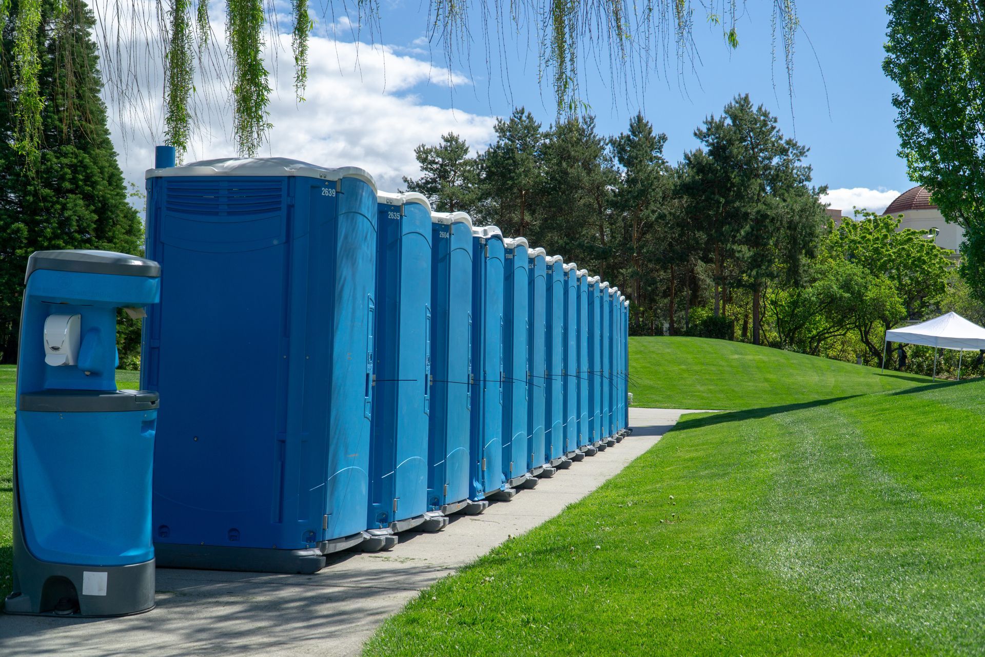 Blue portable toilets lined along a walkway in a landscaped park setting. Blue portable toilets lined along a walkway in a landscaped park setting.