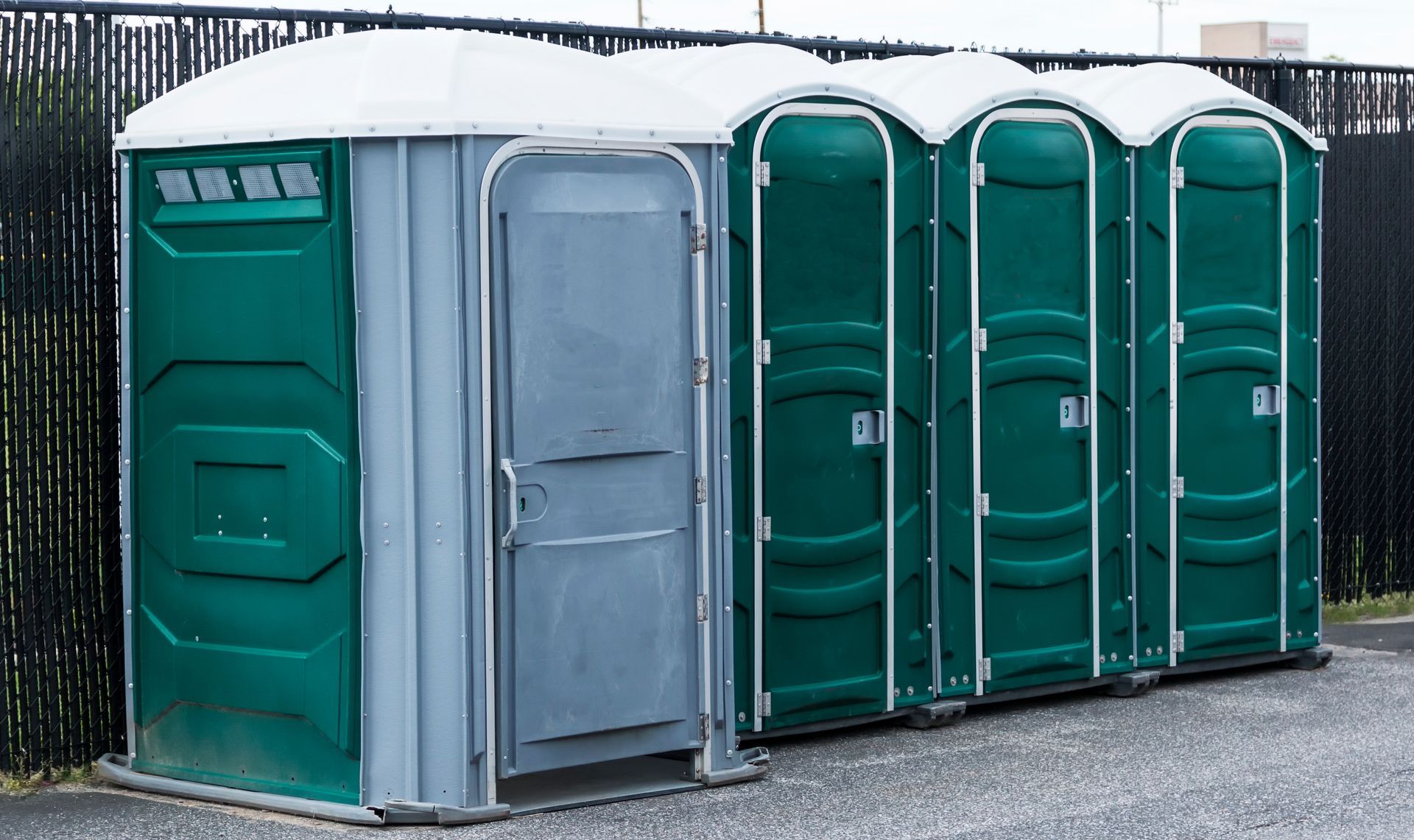 Photograph of four portable toilets lined up outdoors on a paved surface with a black fence in the background. Photograph of four portable toilets lined up outdoors on a paved surface with a black fence in the background.
