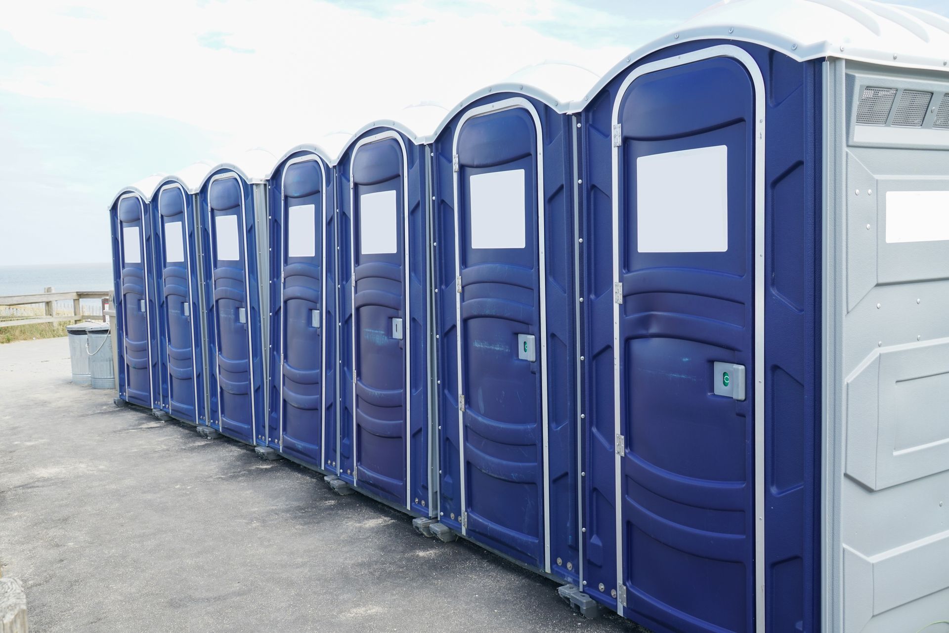 A row of blue portable toilets lined up outdoors on a paved surface near a wooden fence. A row of blue portable toilets lined up outdoors on a paved surface near a wooden fence.