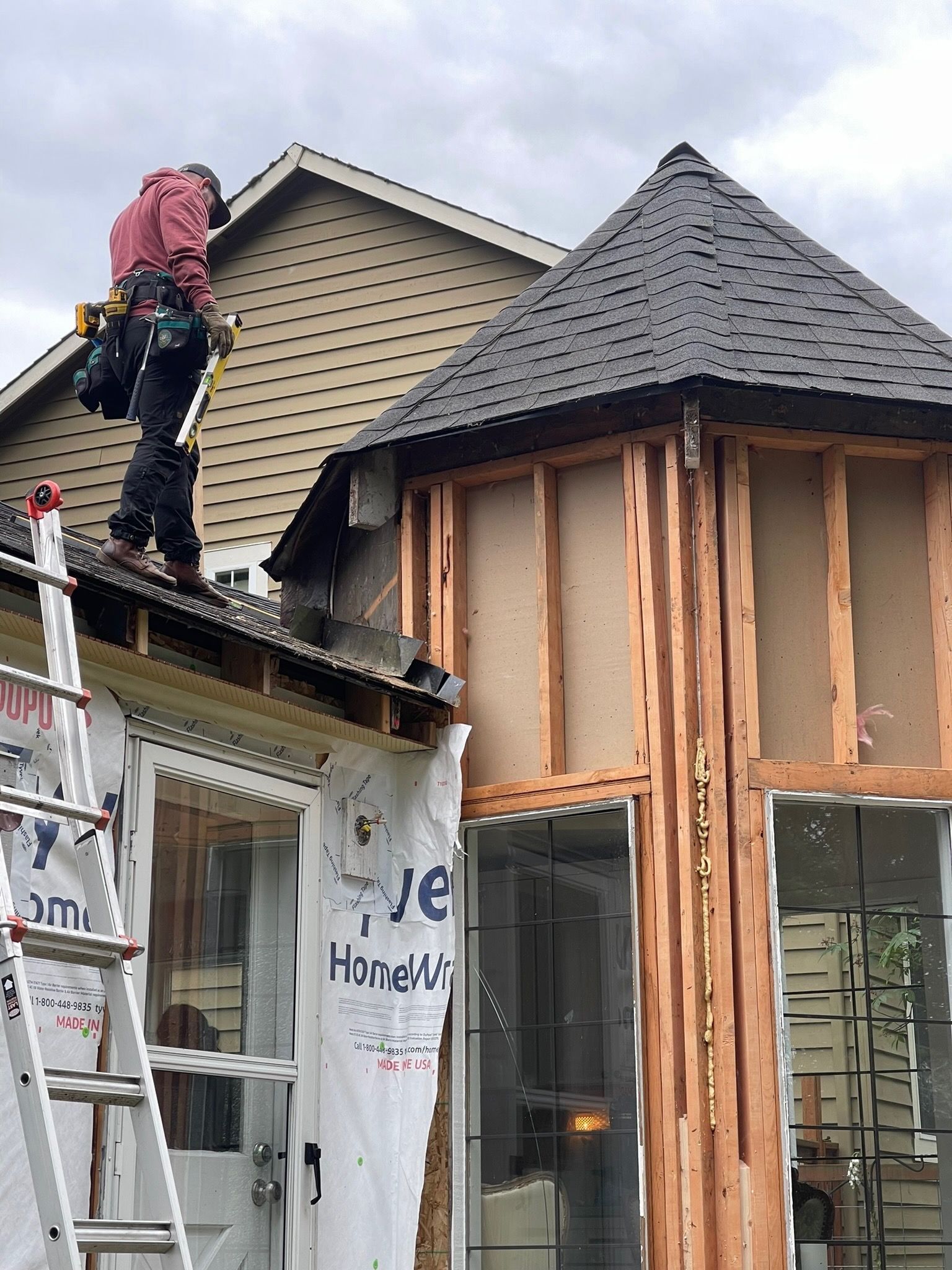 A construction worker in a red shirt stands on a roof next to the unfinished wood framing of a new bay window.