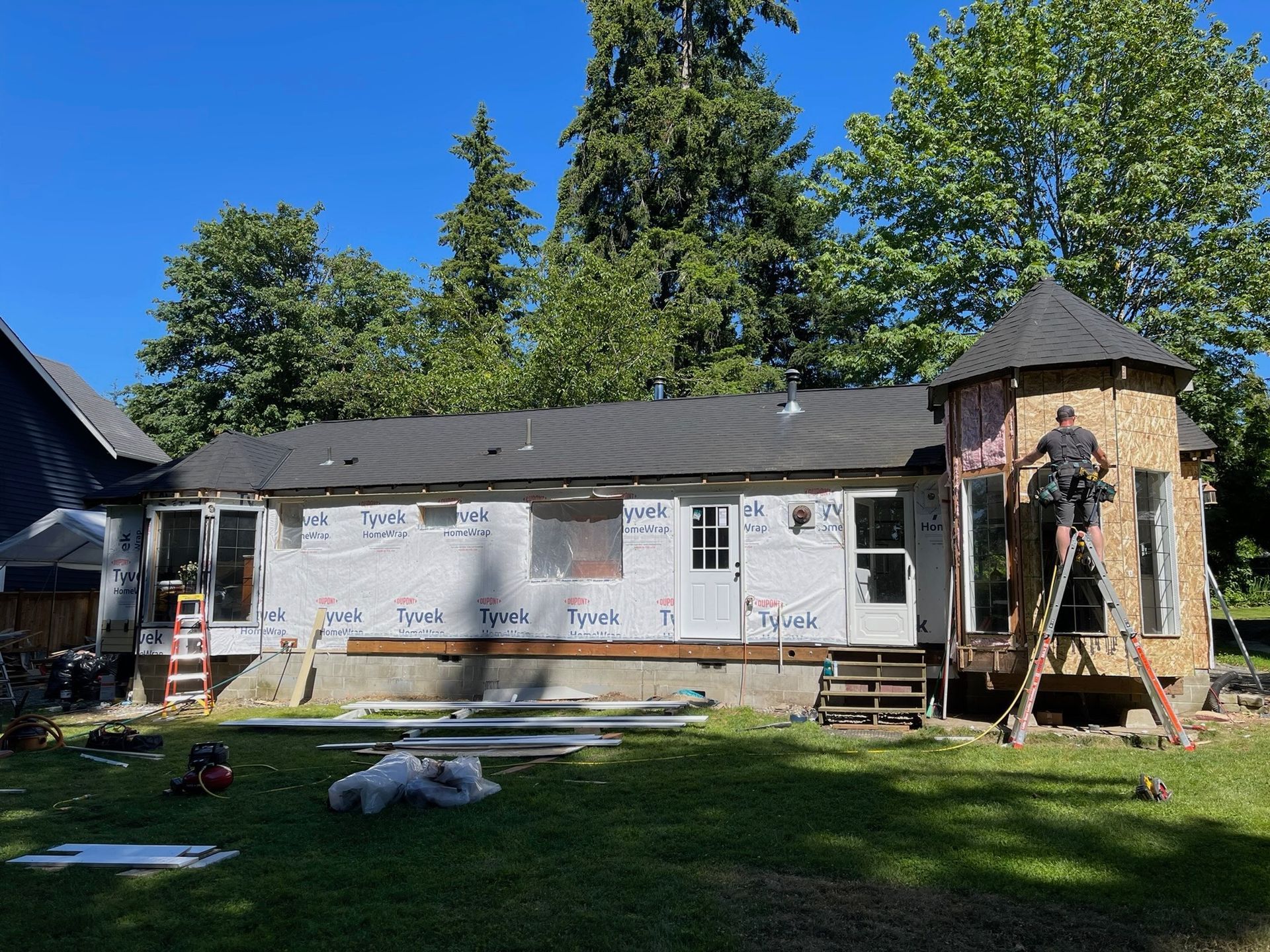 A worker on a ladder installs plywood siding on a turret-style addition to a house under renovation on a sunny day.