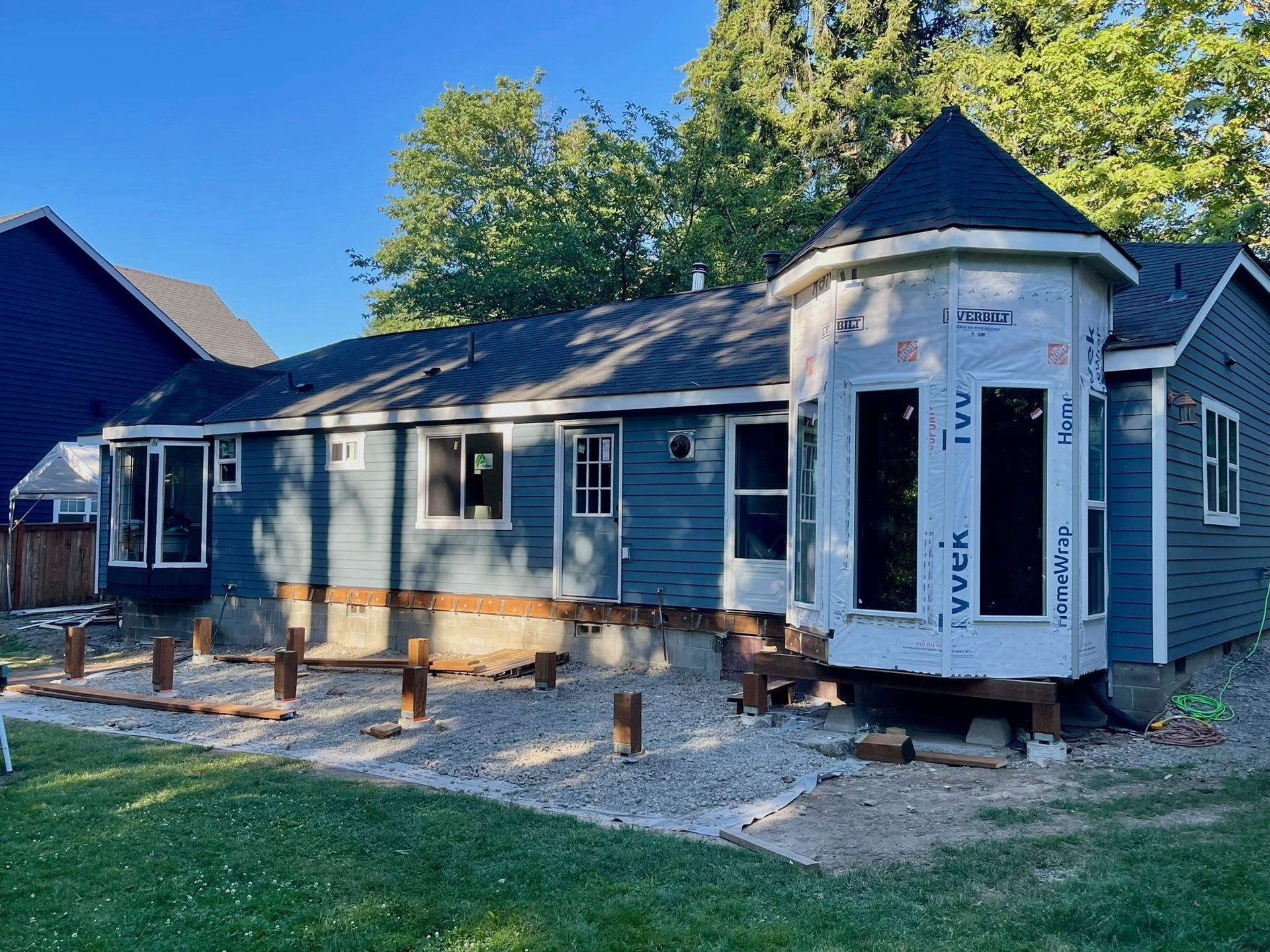 A blue house under construction with exposed foundation, support posts, and a gravel yard.