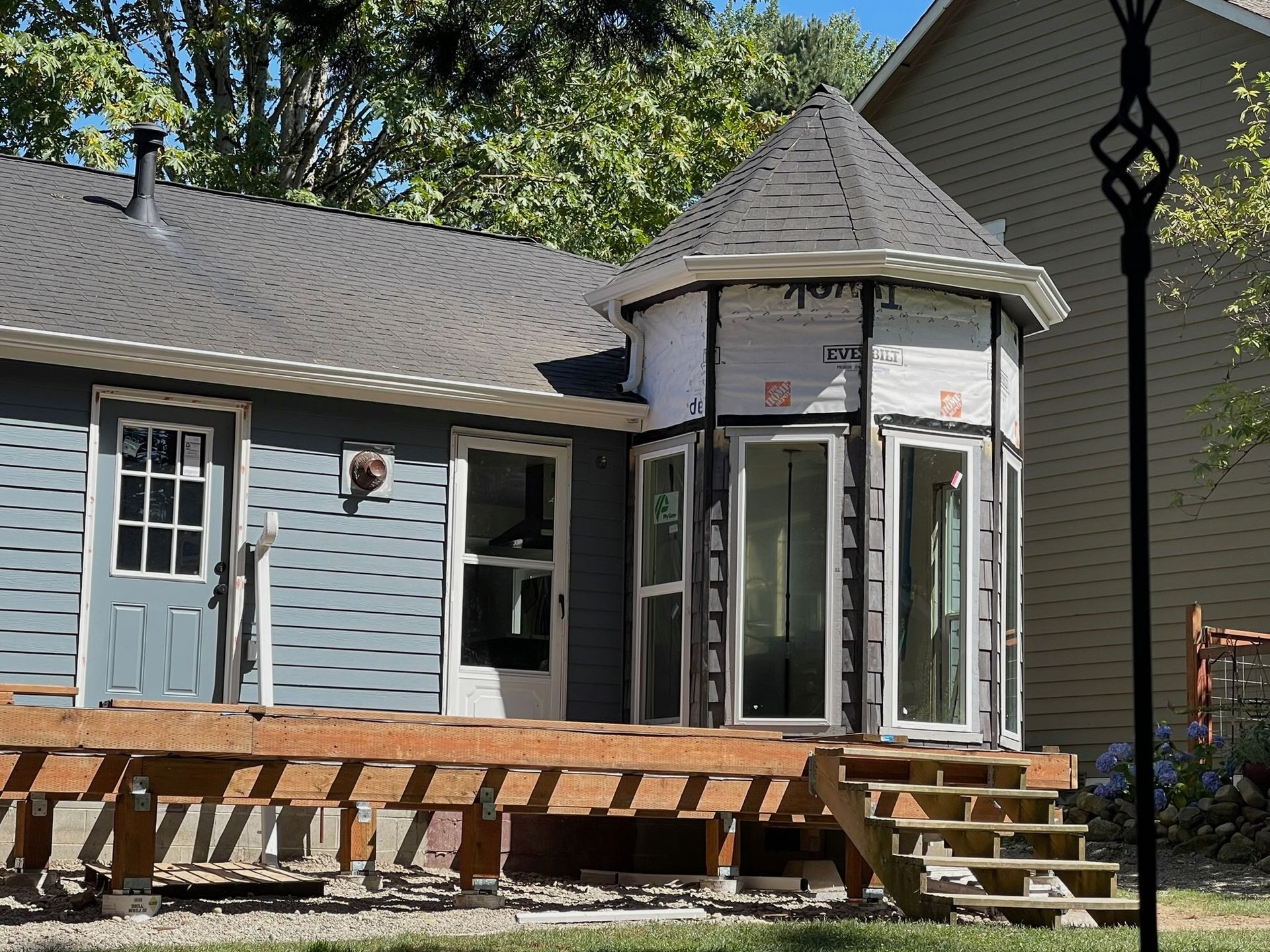 A house exterior featuring a blue-grey wall, a wooden deck under construction, and an unfinished white turret bay window.