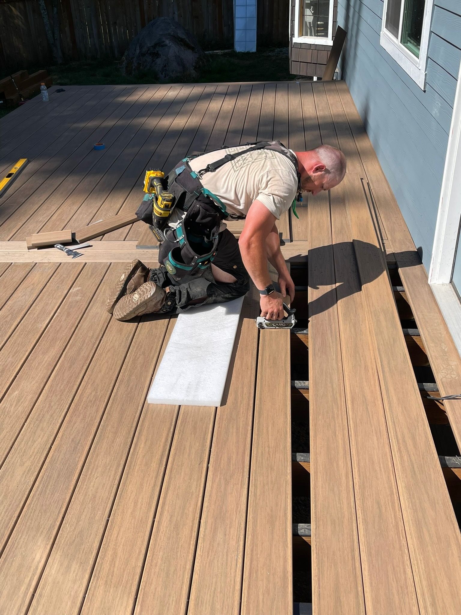 A construction worker kneels on a deck, using a power tool to install brown composite decking boards next to a blue house.