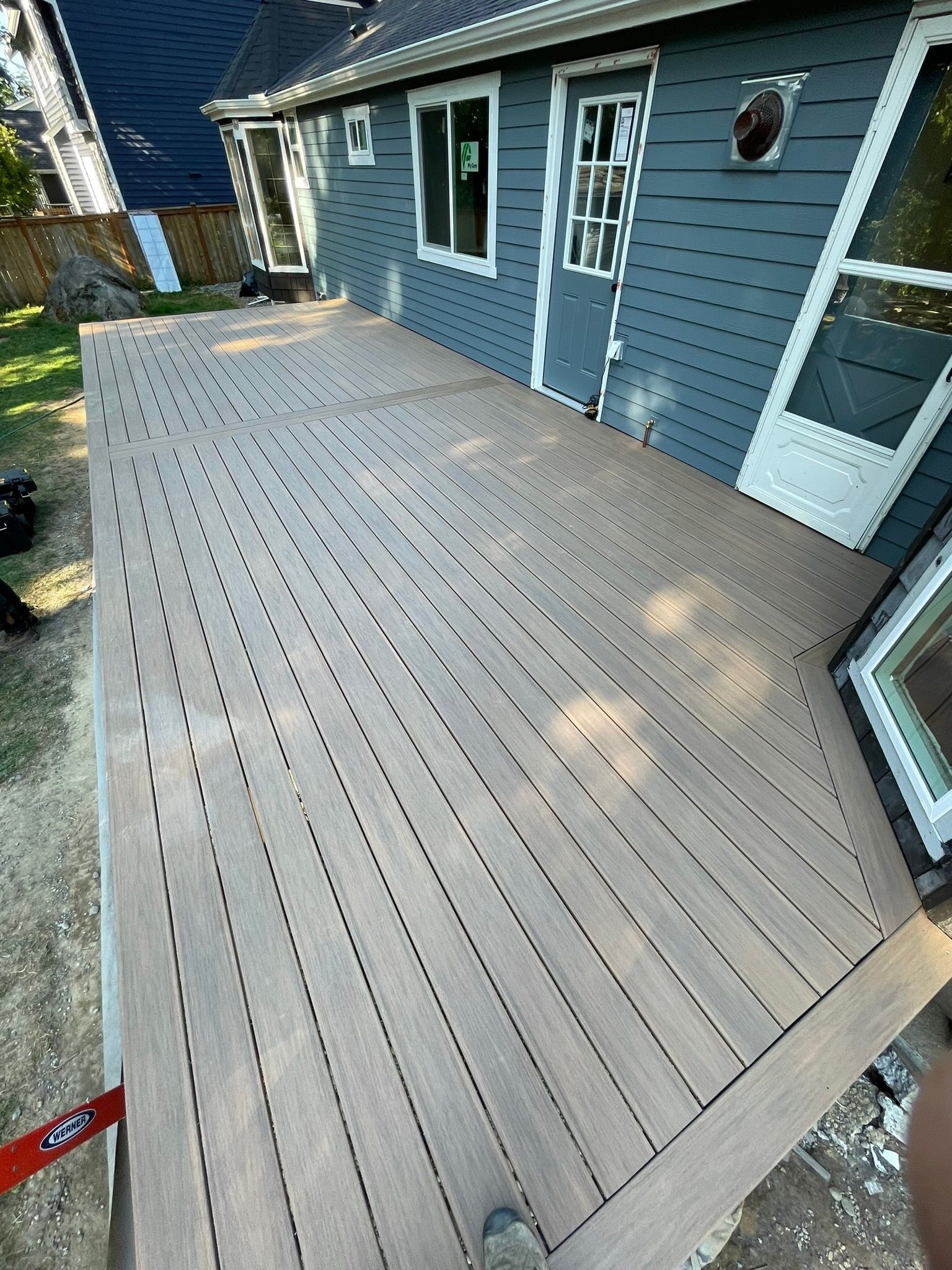 A light-brown composite deck attached to the side of a grey-sided house with white trim.