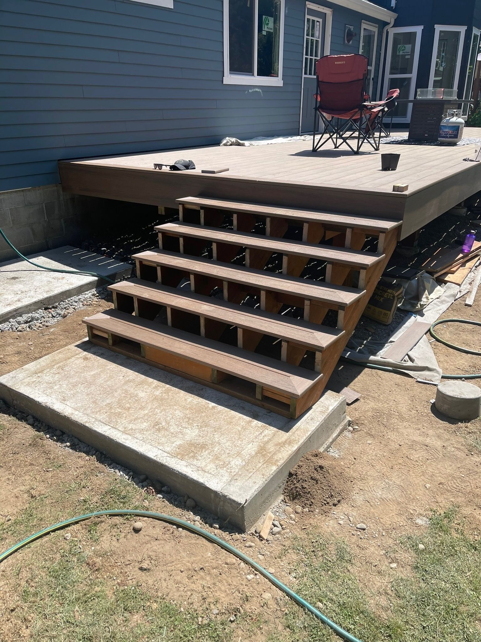 A low-angle view of a brown wooden deck with new steps leading down to a concrete landing at the side of a blue house.