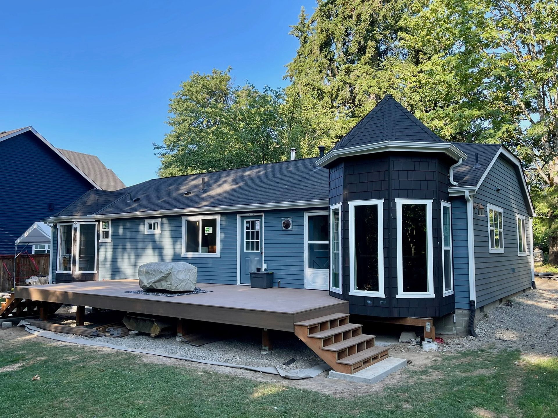 A blue single-story house with a wooden deck, a hexagonal bump-out, and a dark roof in a green, tree-filled backyard.