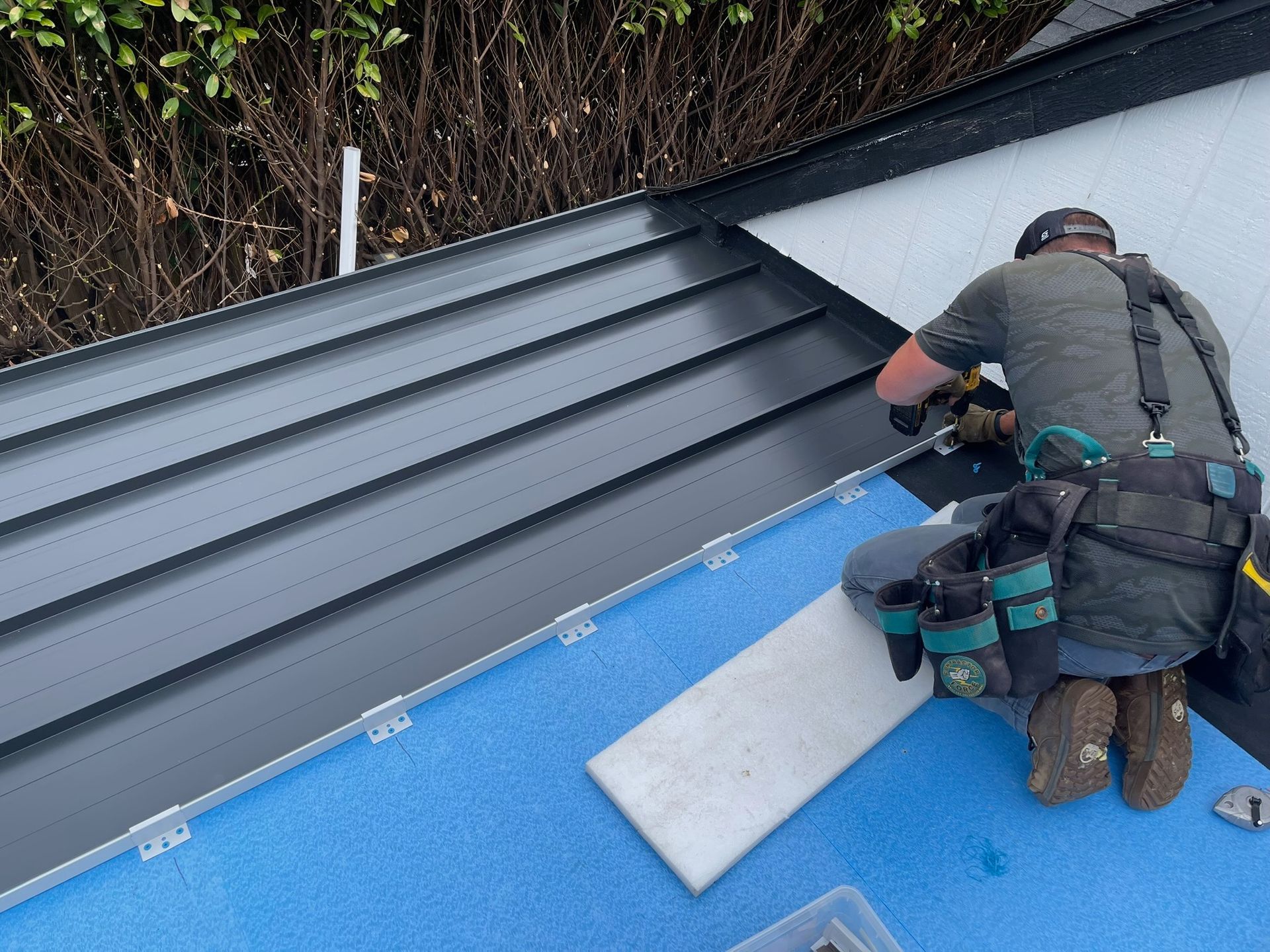 A person in a tool belt installs dark metal roofing panels onto a blue underlayment on a residential roof.