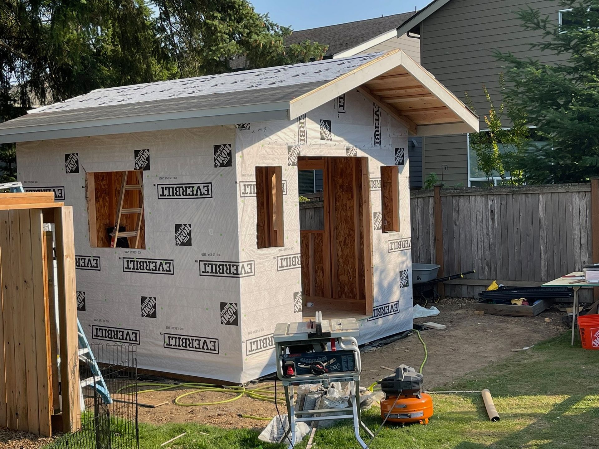 A shed under construction in a backyard, featuring exposed wood framing, white weather-resistant sheathing, and a roof.