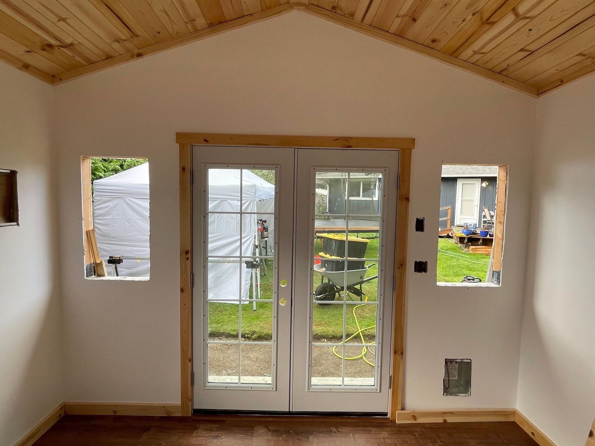 An interior view of a room with white walls, a wood-paneled ceiling, double doors, and two window openings.