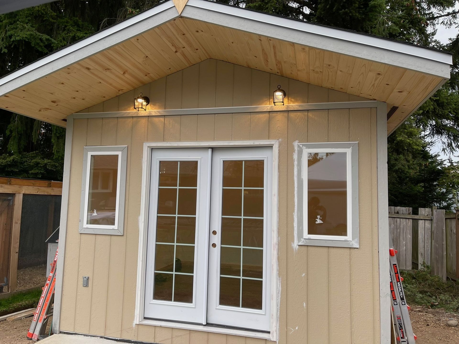 A tan wooden shed with a gabled roof, white-trimmed French doors, and two windows, illuminated by outdoor lights.