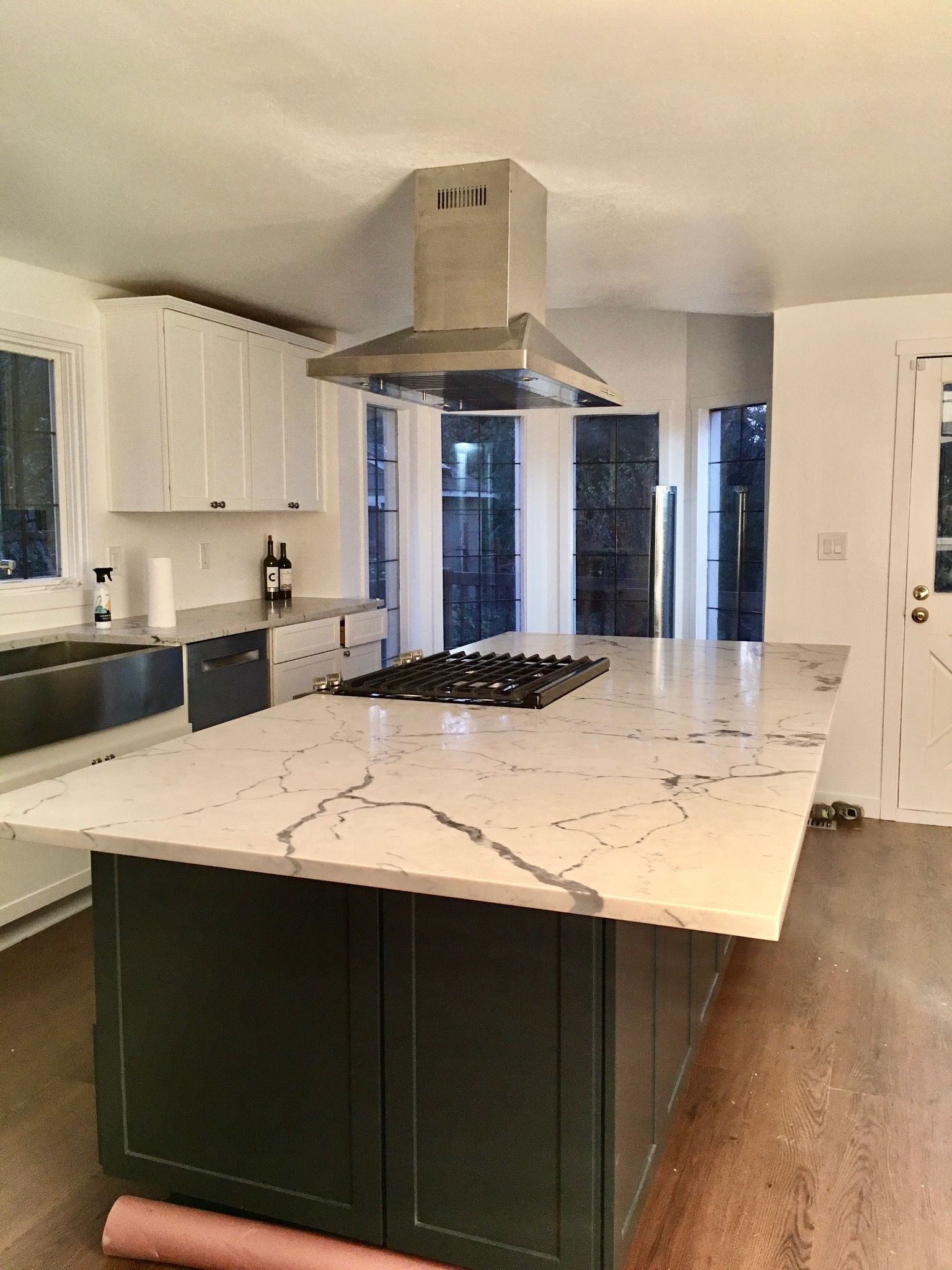 A kitchen island with a white marble countertop and gas cooktop, featuring dark green cabinets and a metal range hood.