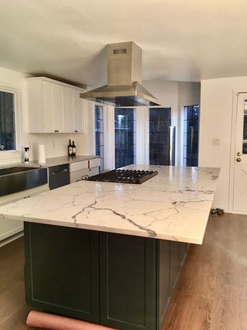 Kitchen island with white marble countertop and cooktop, stainless steel range hood, and dark green cabinetry.