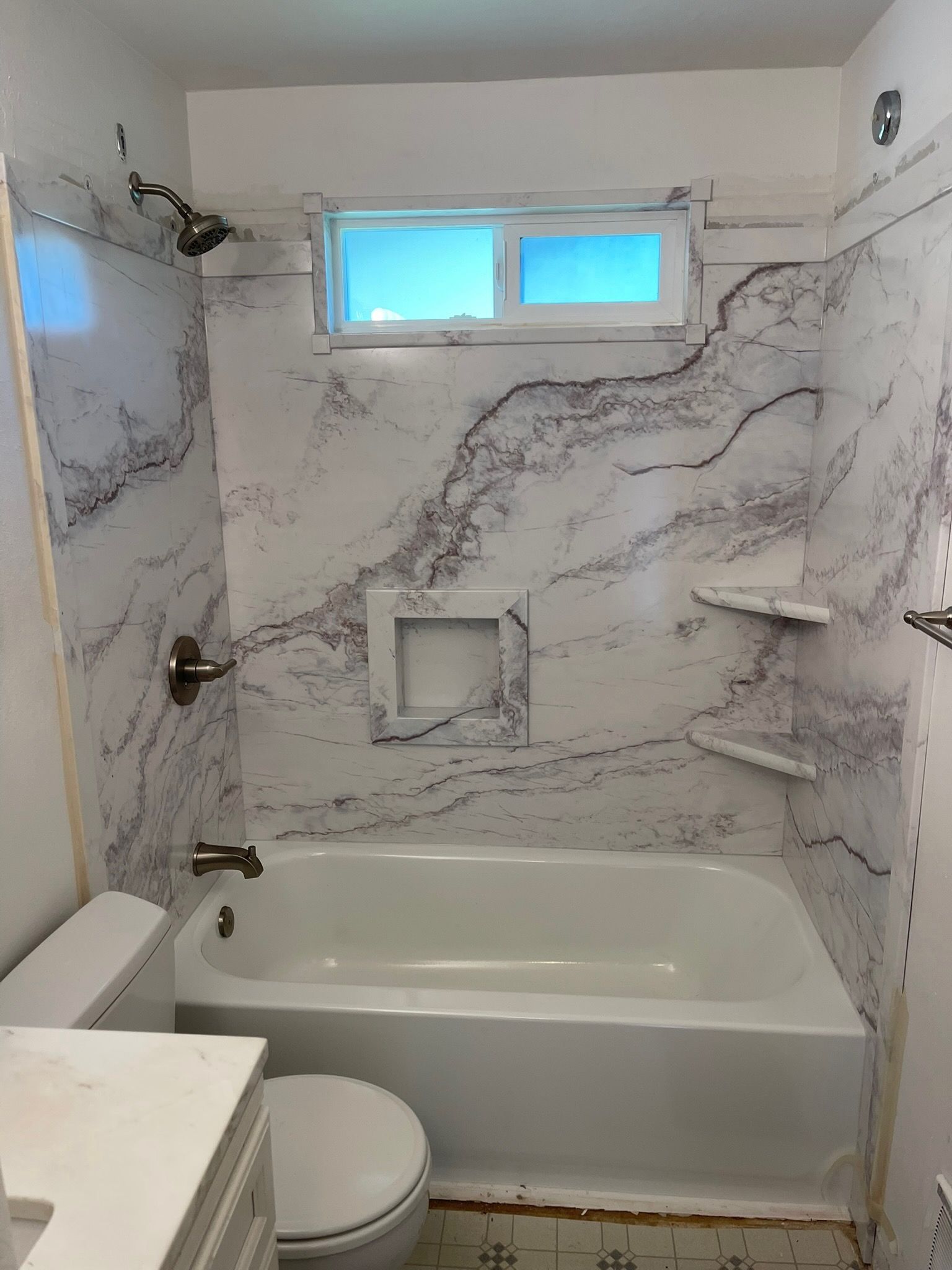A bathtub and shower area with white marble-patterned wall panels, a niche, corner shelves, and chrome fixtures.