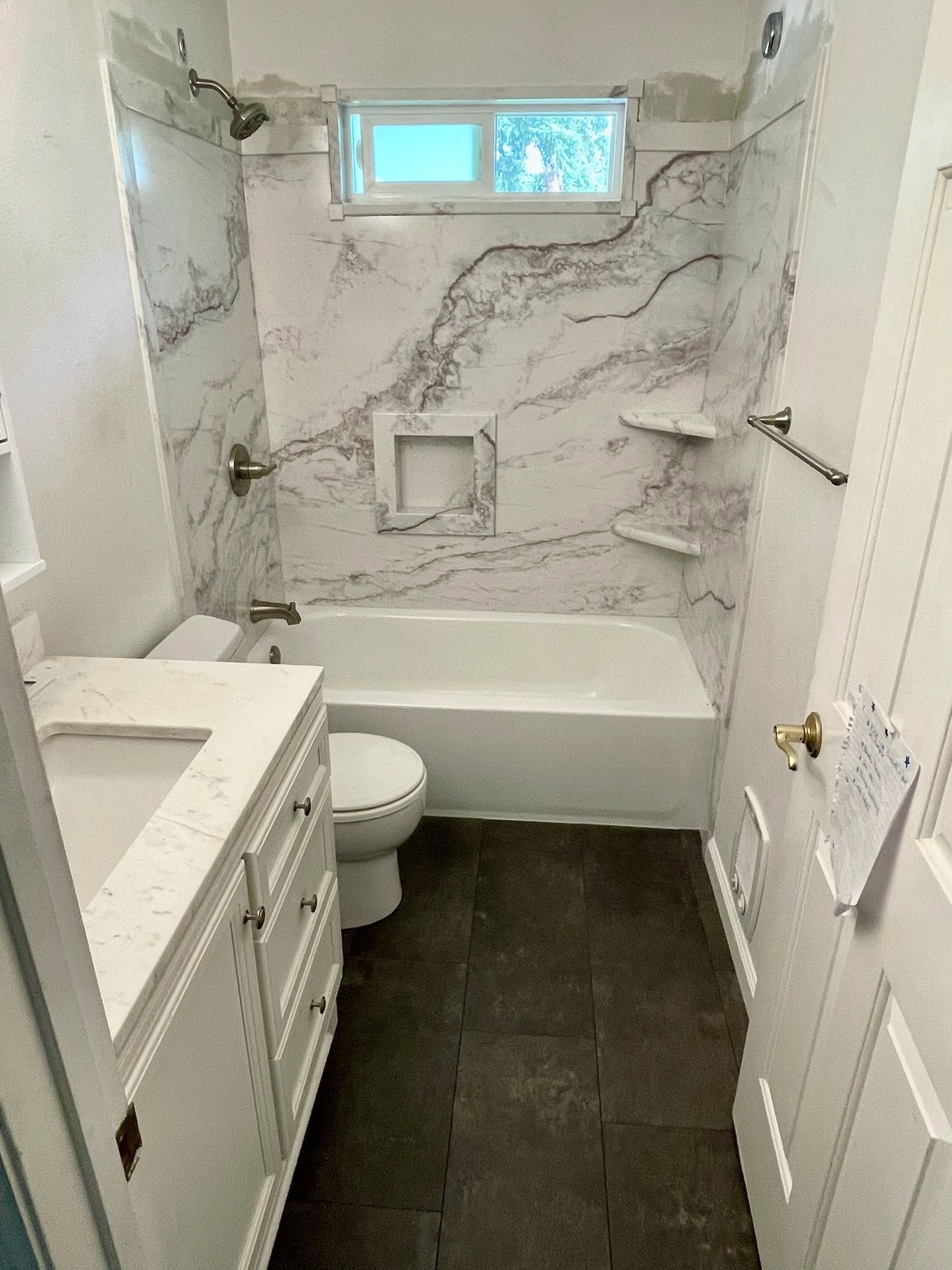 A small bathroom with white cabinets, a white vanity top, a toilet, and a bathtub with marble-patterned tile walls.